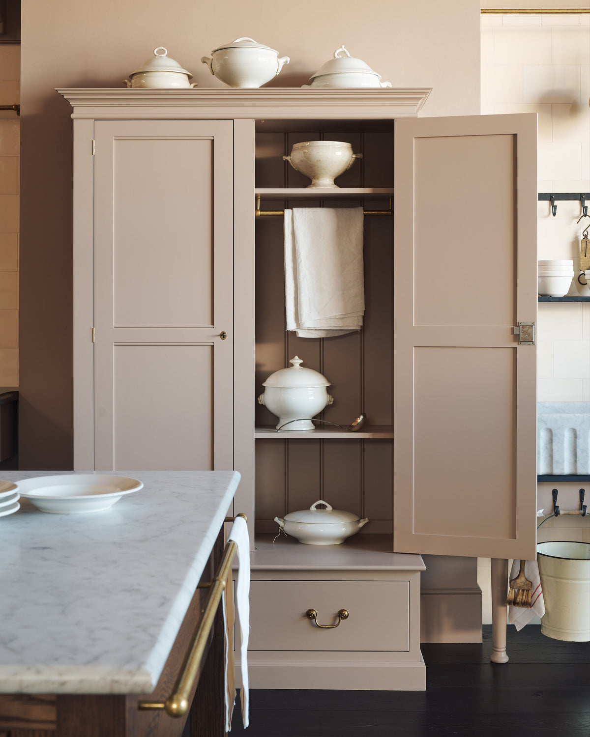 A quiet corner of a grand Princelet pink Heirloom and Classic English kitchen features a sink cabinet with a black granite worktop, a large kitchen island with a Carrara marble worktop, aged brass rails, ditsy delft wallpaper, a tall cream subway tile splashback, and an Heirloom task light. Lovingly adorned with brass pots and pans, an antique painting, and artisan ceramics, creating a warm, delicate, and airy kitchen space.