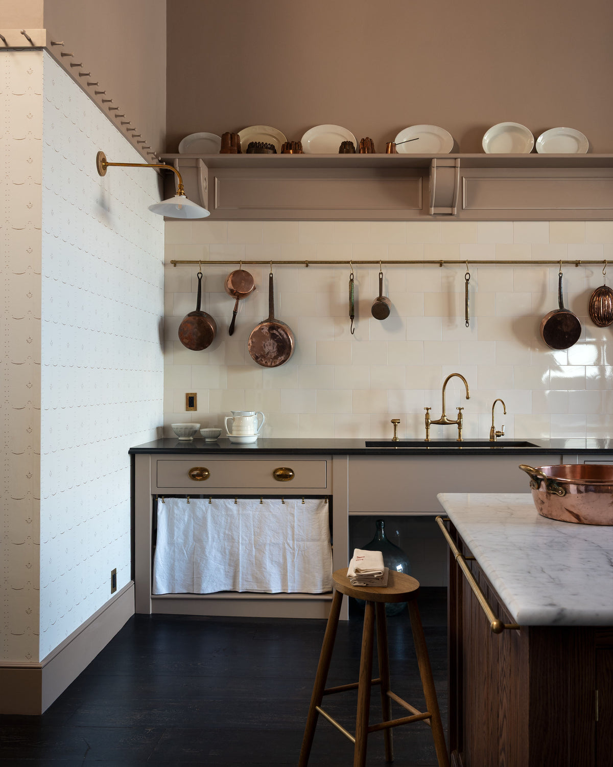 A quiet corner of a grand Princelet pink Heirloom and Classic English kitchen features a sink cabinet with a black granite worktop, a large kitchen island with a Carrara marble worktop, aged brass rails, ditsy delft wallpaper, a tall cream subway tile splashback, and an Heirloom task light. Lovingly adorned with brass pots and pans, an antique painting, and artisan ceramics, creating a warm, delicate, and airy kitchen space.
