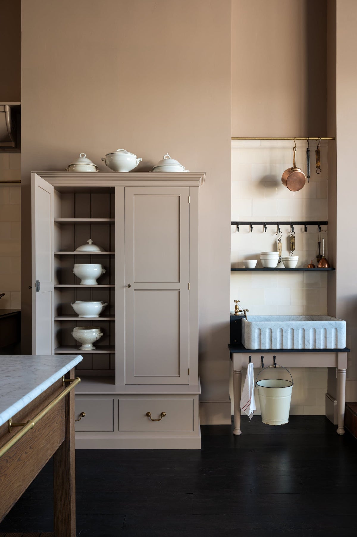 A quiet corner of a grand Princelet pink Heirloom and Classic English kitchen features a sink cabinet with a black granite worktop, a large kitchen island with a Carrara marble worktop, aged brass rails, ditsy delft wallpaper, a tall cream subway tile splashback, and an Heirloom task light. Lovingly adorned with brass pots and pans, an antique painting, and artisan ceramics, creating a warm, delicate, and airy kitchen space.