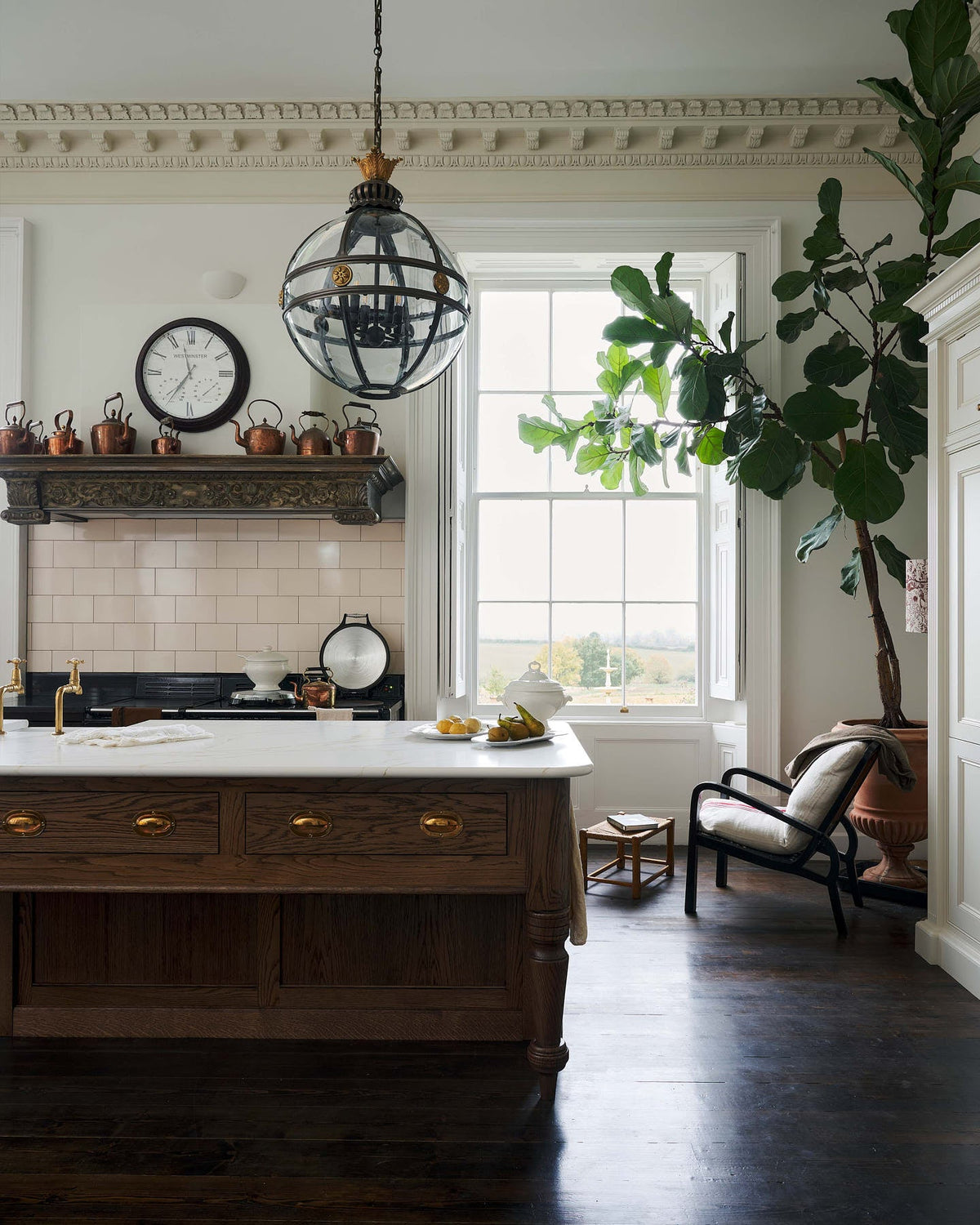 A quiet corner of a simple yet grand Heirloom kitchen features a bespoke 3.5-metre-long dairy table with a single fluted Belfast sink, aged brass Mayan taps, a dishwasher, and a breakfast bar, creating a timeless and practical space. It is decorated with a vintage over mantle, artisan ceramics, glass and metal pendant lights, copper pans, and a large fiddle leaf fig plant.