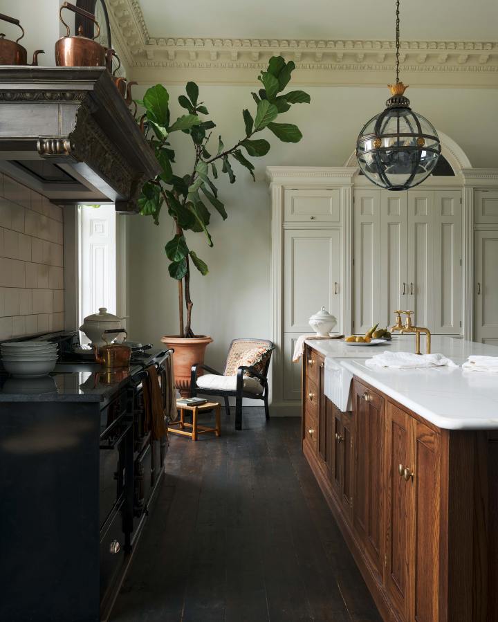 A simple yet grand Heirloom kitchen features a bespoke 3.5-metre-long dairy table with a single fluted Belfast sink, aged brass Mayan taps, a dishwasher, and a breakfast bar, creating a timeless and practical space. It is decorated with a vintage overmantle, artisan ceramics, glass and metal pendant lights, copper pans, and a large fiddle leaf fig plant.