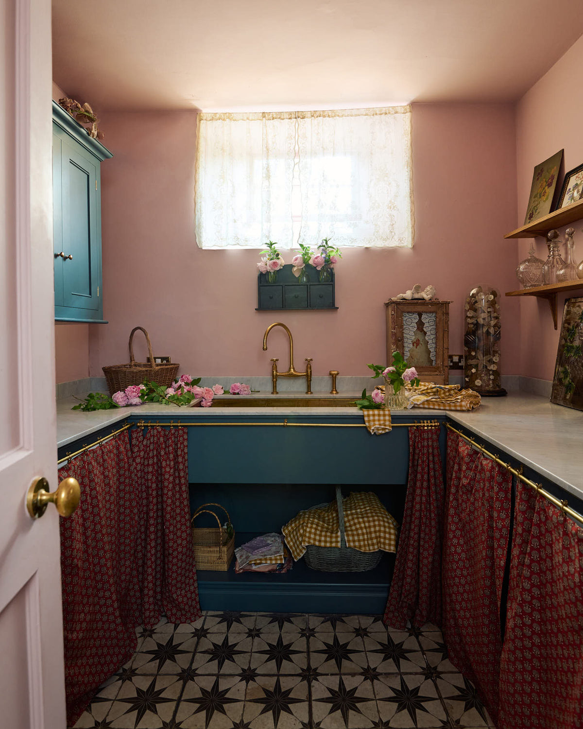Past a blush pink door is a small and practical Heirloom laundry room featuring a sink cabinet, a polished brass sink, an aged brass Ionian tap, a curtain rail with crest-patterned red curtains, wall cupboards, and shelves. Adorned with eclectic vintage items, pink peonies, and a small wicker basket, this space creates a romantic and functional atmosphere full of personality.