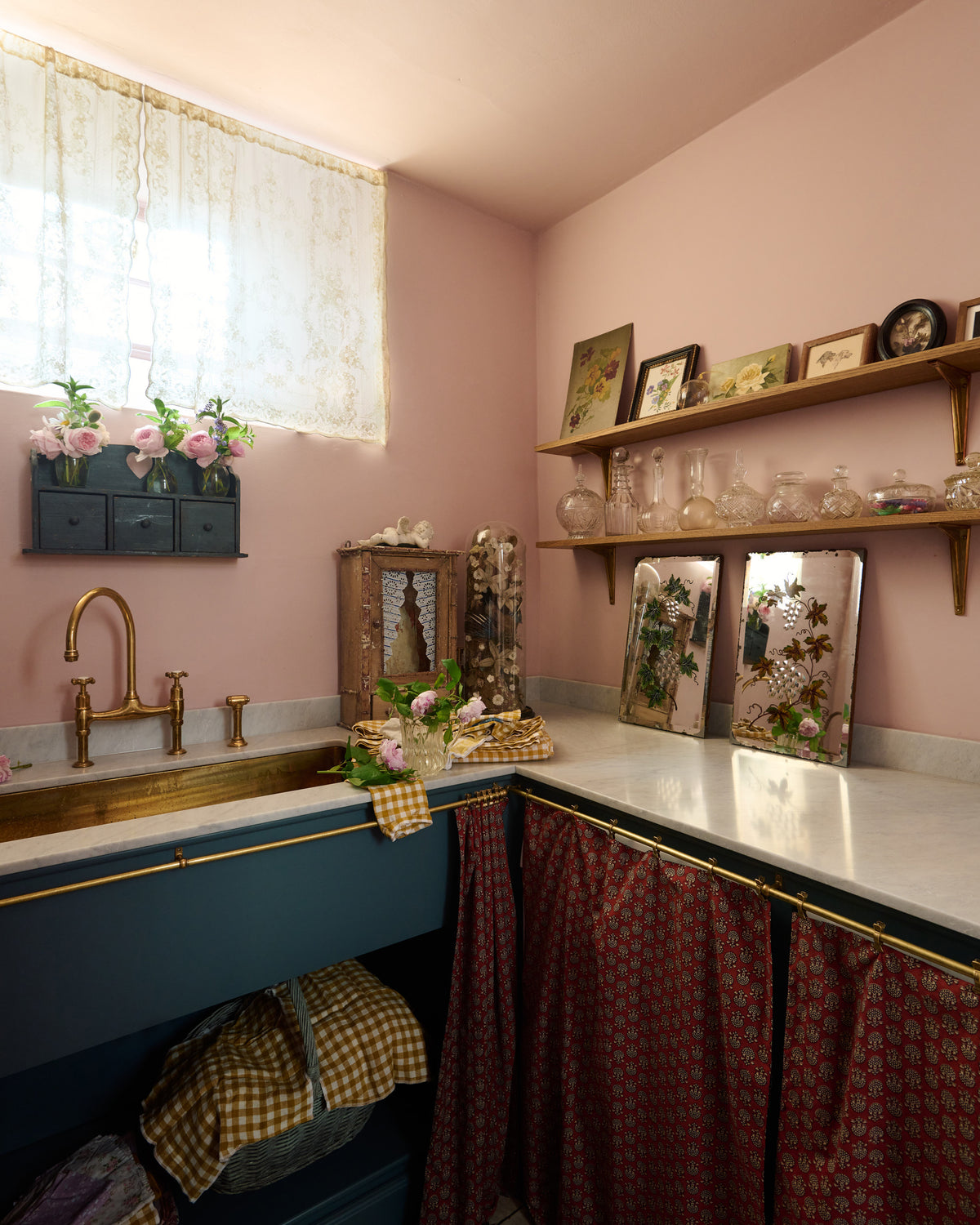 An oblique view of a small and practical Heirloom laundry room features a sink cabinet, a polished brass sink, an aged brass Ionian tap, a curtain rail with crest-patterned red curtains, and shelves. Adorned with eclectic vintage items, pink peonies, tea towels, glass containers, and wicker baskets, this space creates a romantic and functional atmosphere full of personality.