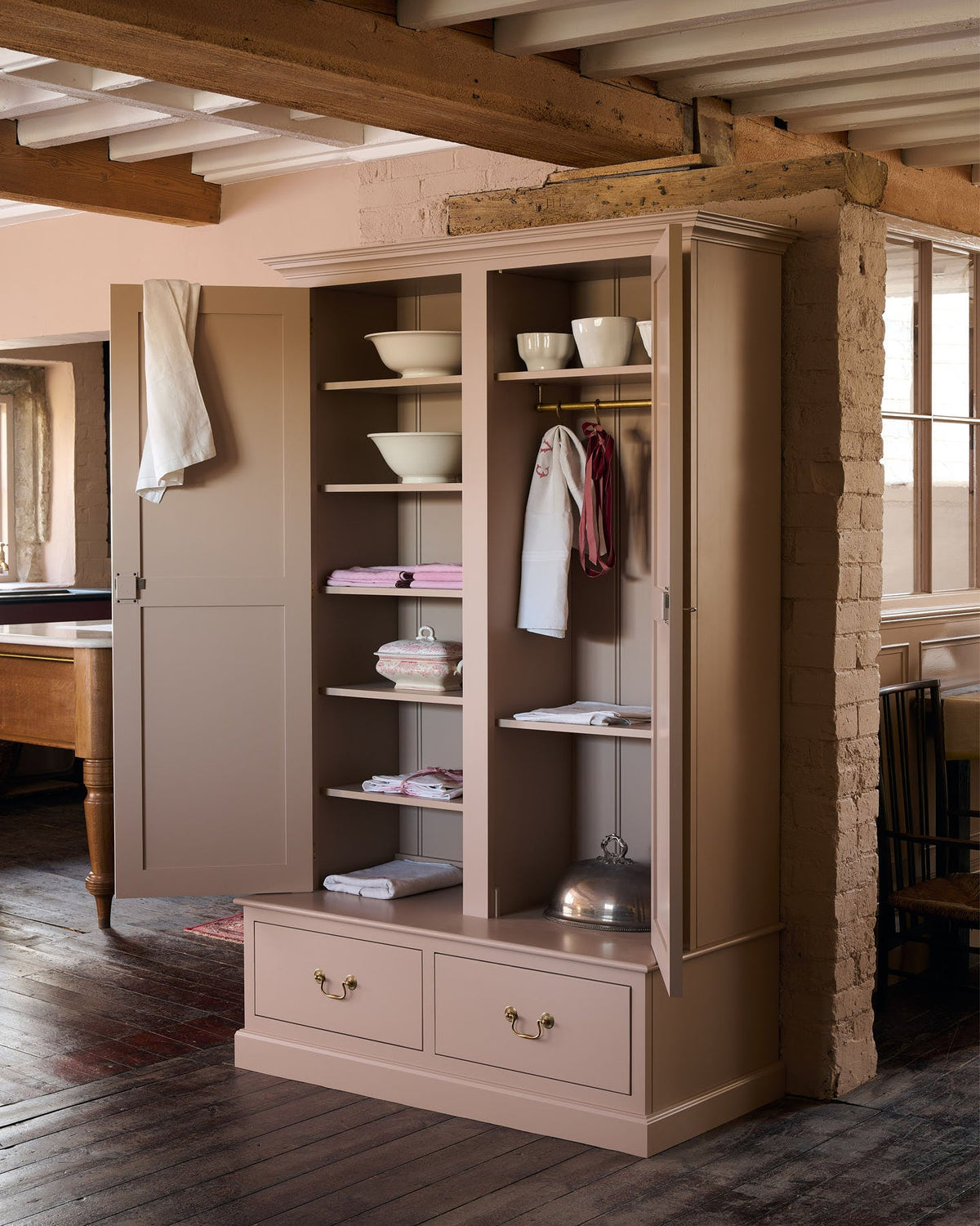 An oblique view of a Victorian-style pantry cupboard, painted inside and out in a soft pink colour. Both doors are open, revealing fixed shelving on the left and a tall hanging space on the right. A pedestal base contains two box drawers with Georgian-style brass pull handles.