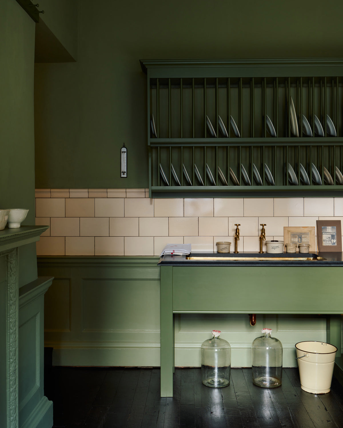 A quaint and stylish Wilkes green Heirloom kitchen features a sink cabinet with a café rail, white lace curtains, a polished brass sink, aged brass Mayan taps, and a large plate rack. Adorned with vintage photos, a cream bucket, simple ceramics, cream subway splashback tiles, and a golden-framed sliced green pumpkin, this kitchen creates a luxurious dining and social space.