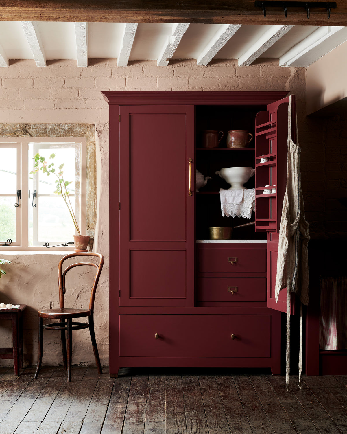 A quiet corner of a refectory-red Heirloom kitchen features a pantry cupboard fitted with brass details featuring pull handles, round knobs, an external drawer, and card frame pulls on internal drawers.