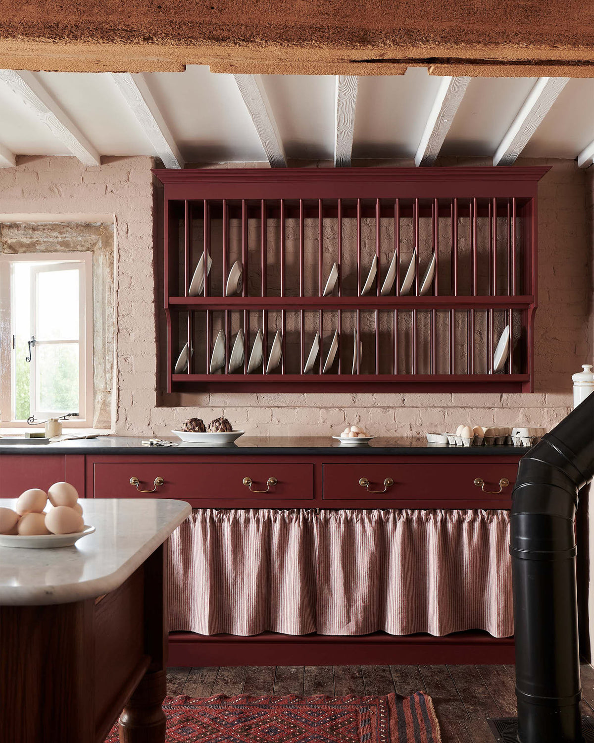 A refectory-red Heirloom kitchen’s sink cabinet reveals a polished brass sink, aged brass Mayan taps, café curtain rails, a red and white striped curtain, a plate rack, and an Heirloom dairy table. Beautifully decorated with artisan ceramics and plates of fresh eggs, this kitchen offers a modern twist on vintage furniture, creating a classic range designed to last.