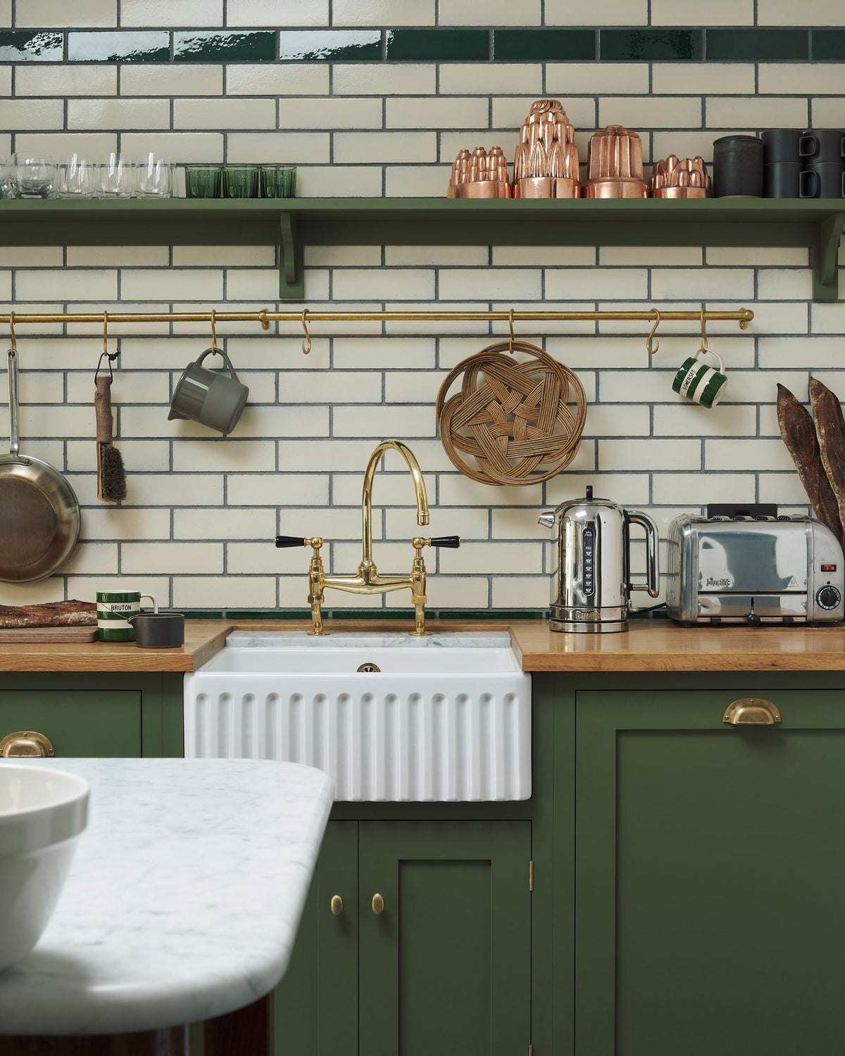A closer look at the quaint and modern Wilkes green Shaker kitchen features a cooker and sink run with a rustic oak worktop, a long shaker shelf, and a Dairy table with a breakfast nook. Adorned with a long hanging rail, signature ceramics, brass cups, and a large wall of metro tiles it embodies a playful industrial and practical design.