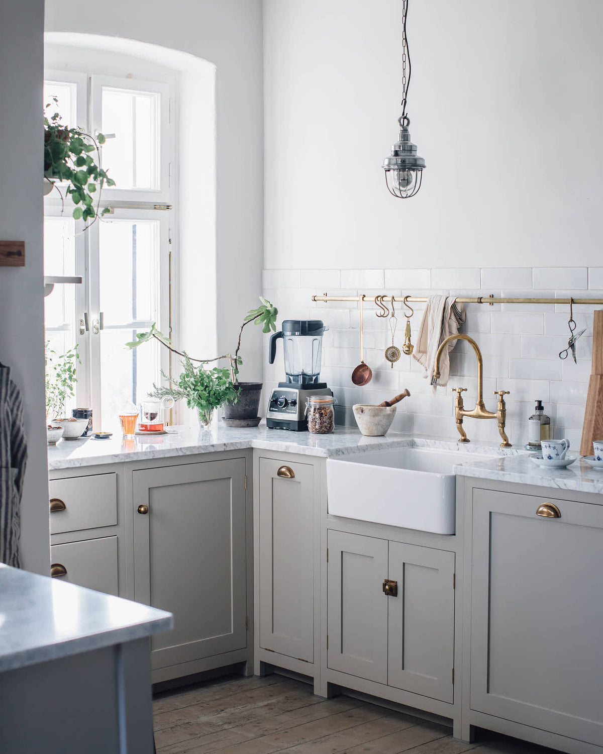 An angled corner of the humble Mushroom colour shaker kitchen with an all-in-one L-shaped sink and cooker run, and a small rolling kitchen island with Carrara marble worktop. A single Belfast sink is complemented by an aged brass Ionian tap, and an aged brass hanging rail adds a striking element against the grey and neutral tones of this charming kitchen.