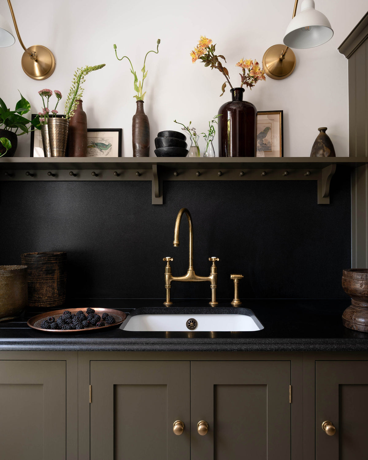 A closer look at the murky-green galley shaker kitchen, featuring a sink run with a black granite worktop and splashback, an undermounted sink, an aged brass ionian tap, and a shaker shelf with pegs. Beautifully curated with two brass wall lights, artisan ceramics, fresh houseplants, framed vintage illustrations, and a brass plate of blackberries, it creates a chic, understated, and practical galley kitchen.