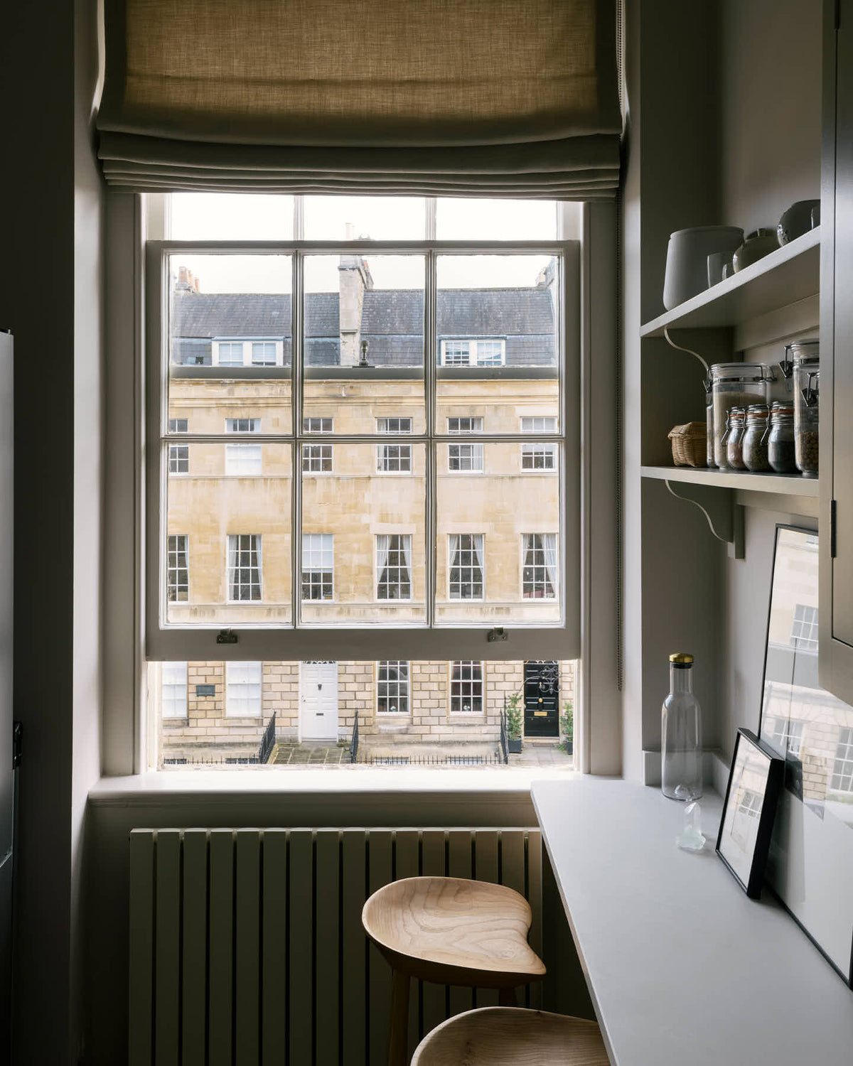 A stylish, mushroom-coloured Shaker galley kitchen features a cooker and sink run with Silestone worktop and a cosy breakfast nook. Beautifully decorated with simple, everyday kitchen items such as dried ingredients, muted ceramics, framed illustrations, deVOL Bum Chairs, a healing crystal, and an amazing view of Bath’s famous Georgian apartments, this calming and functional kitchen space blends character with tranquillity.
