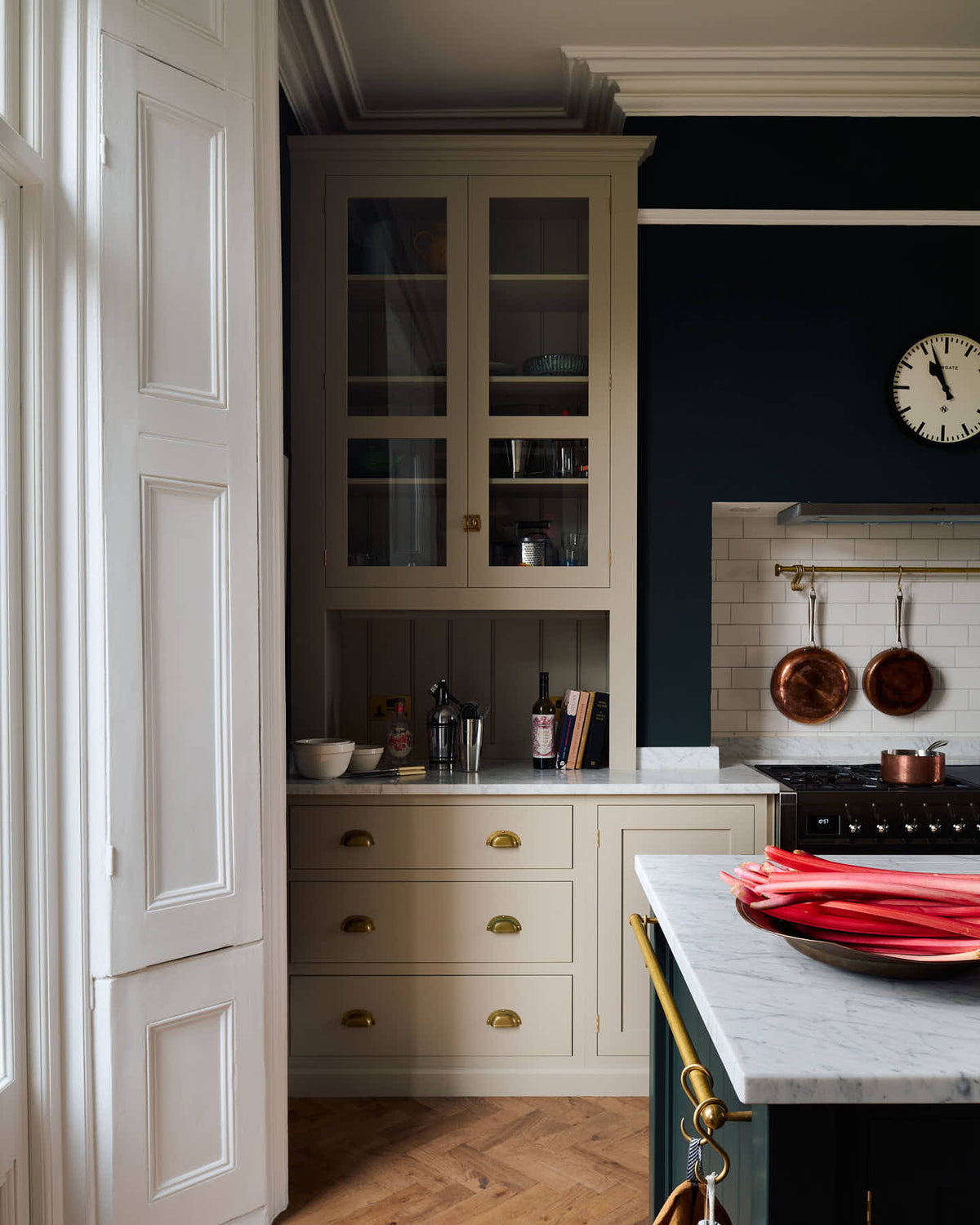 A quiet corner of a simple and elegant mushroom-brown Shaker kitchen features a cooker run with freestanding cupboards, a Carrara marble worktop, a false chimney breast, white porcelain splash back tiles, and a large kitchen island. The space feels loving and functional, adorned with artisan ceramics, copper pans, bright red rhubarb, luscious houseplants, and books.