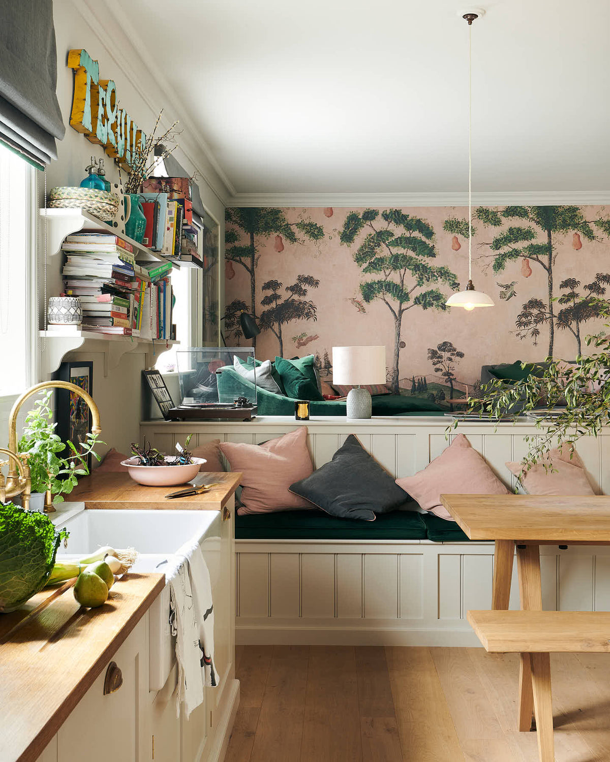 A vibrant Shaker kitchen features an L-shaped sink and cooker run with a solid oak worktop, a single Belfast sink, an aged brass Ionian tap, a Shaker shelf, and a bench. Lovingly decorated with pink rainforest wallpaper, colourful cushions, overstocked books, a record player, chattered skullcap porcelain lights, and a park bench dining table, it creates a characterful and inviting space.