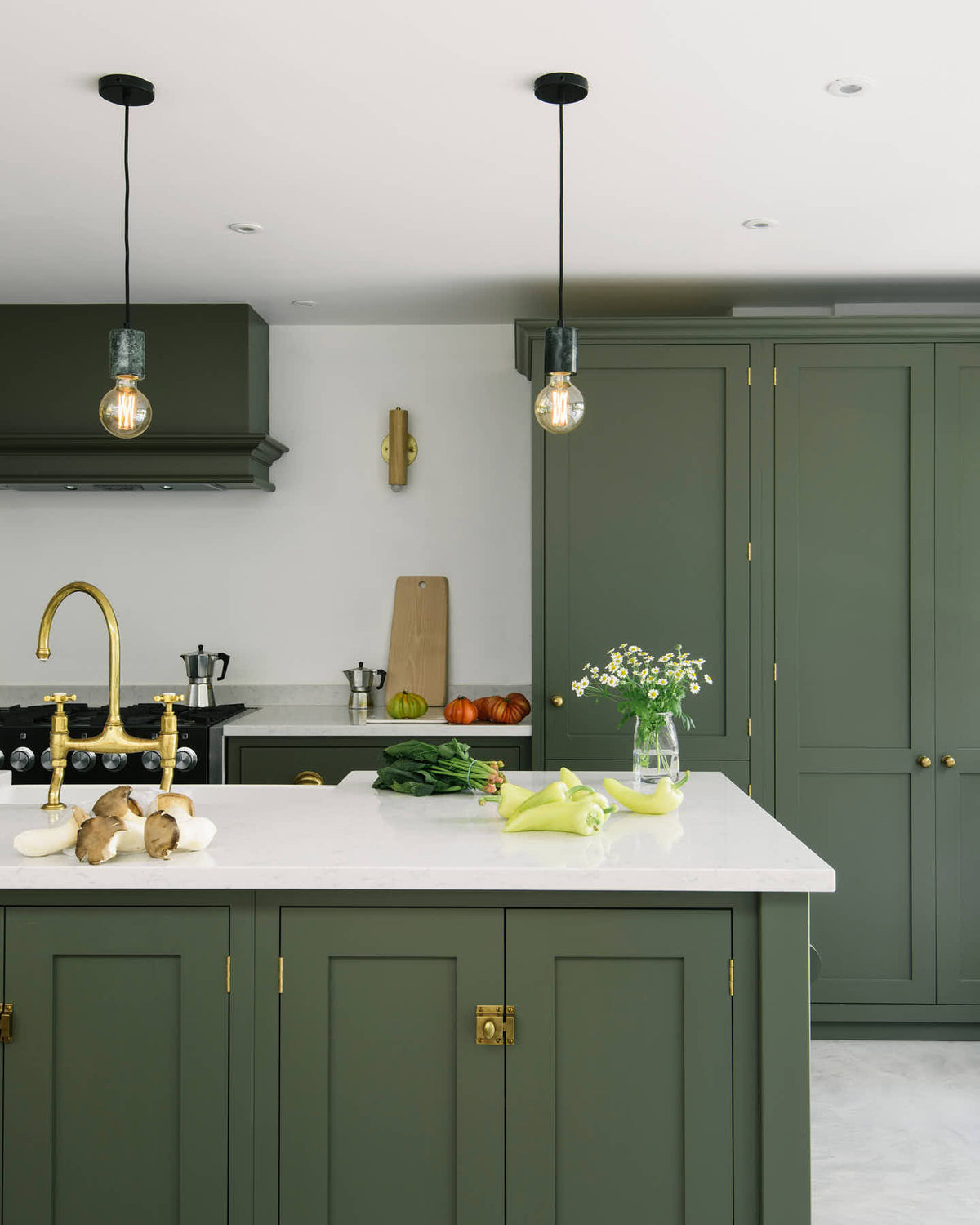 A bright and sophisticated Bakehouse Green shaker kitchen, styled with Scandinavian furniture and a soothing green palette. The kitchen features a cooker run, a central island with a double Belfast sink and silestone worktop, and full-height cupboards. Natural light floods the kitchen and dining room through the glass French doors.