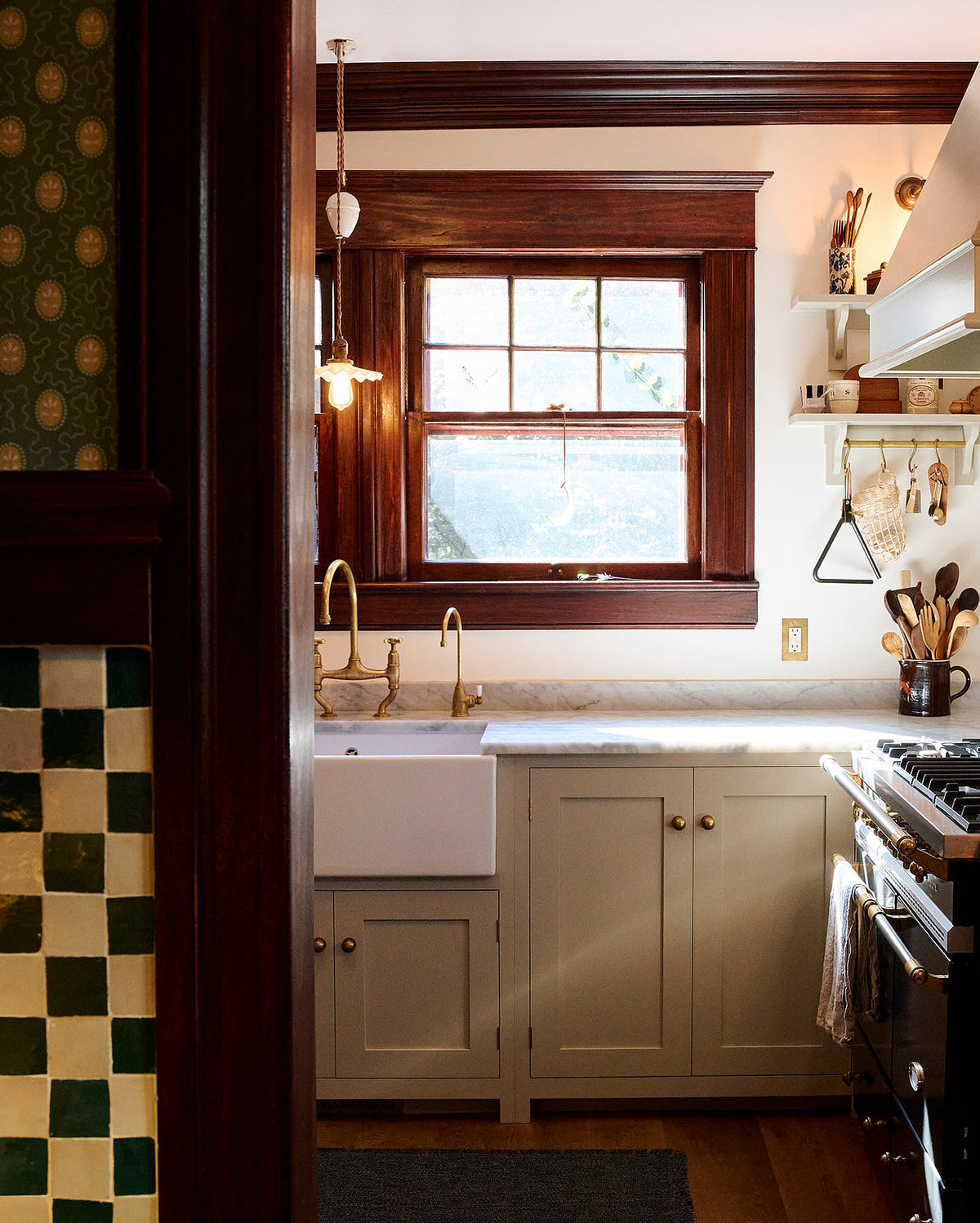 Peeking through a green-and-white mosaic wall and moss-green wallpaper is a humble mushroom-painted shaker kitchen. It features a single Belfast sink with an aged brass Ionian tap and a Carrara marble worktop. A rich brown-framed window floods the space with natural light and serenity.