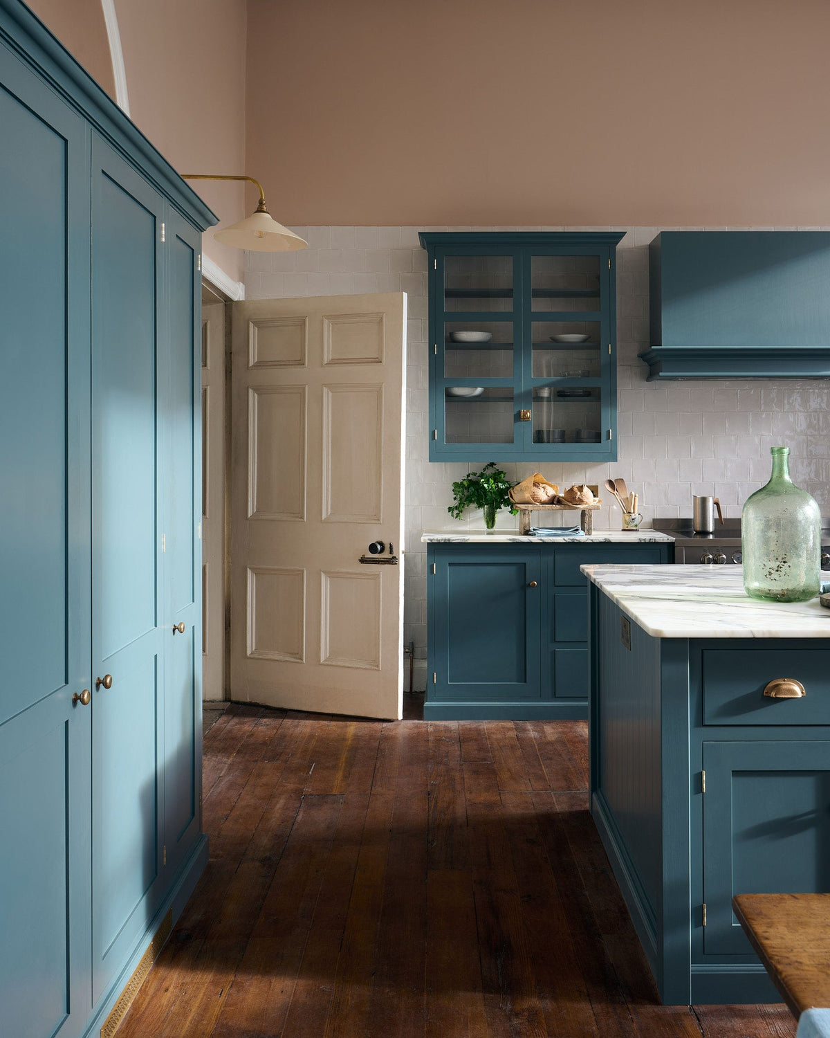 The room, a grand Georgian space from the 18th century, features charming pink walls. It showcases a modern Clerkenwell Blue shaker kitchen equipped with glazed wall cupboards, a shaker-style canopy, a cooker run, white porcelain splash back, large full-height pantry cupboards, and a spacious kitchen island with a sink and an Arabescato marble worktop.