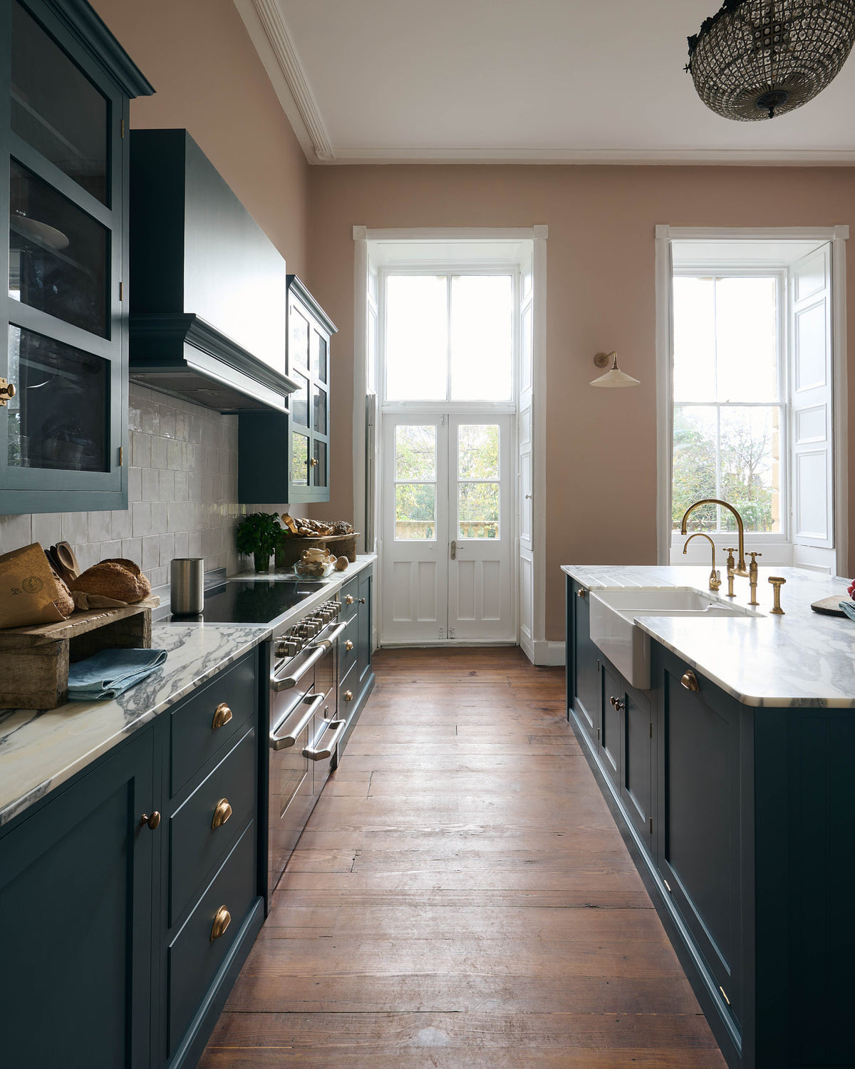 The room, a grand Georgian space from the 18th century, features charming pink walls. It showcases a modern Clerkenwell Blue shaker kitchen equipped with glazed wall cupboards, a shaker-style canopy, a cooker run, white porcelain splash back, large full-height pantry cupboards, and a spacious kitchen island with a sink and an Arabescato marble worktop.