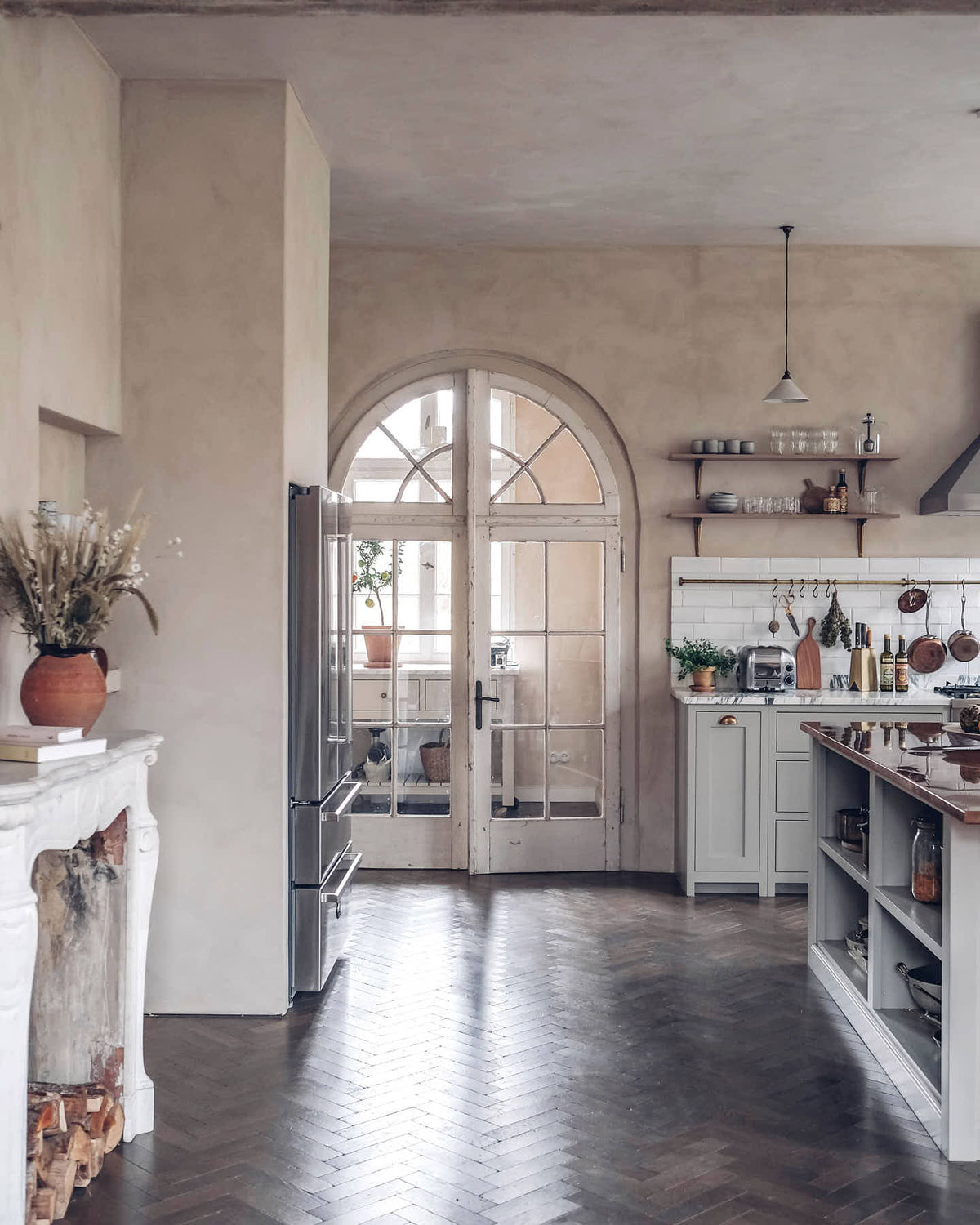 Showing a corner of a grand yet grounded shaker kitchen in muted mushroom off-white, alive with scattered pots, pans, fruits, and vegetables. A large copper-topped island and cosy breakfast nook complete the space. Elegant arched glass doors open to a small hallway with a freestanding table, highlighting the movement of rich oak parquet floors.