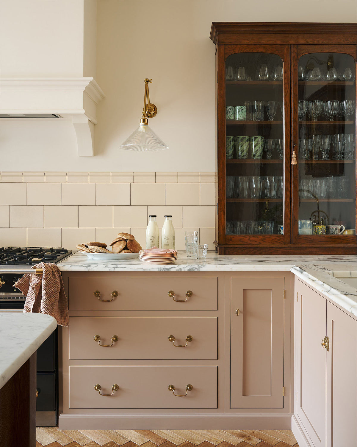 A beautiful corner of a soft Princelet pink Shaker kitchen unfolds with an L-shaped cooker and sink run topped in Carrara marble. It features a counter cupboard, cream subway tiles, and a large Dairy Table. The kitchen is decorated with milk, glasses, and pitta breads in a large ceramic dish.