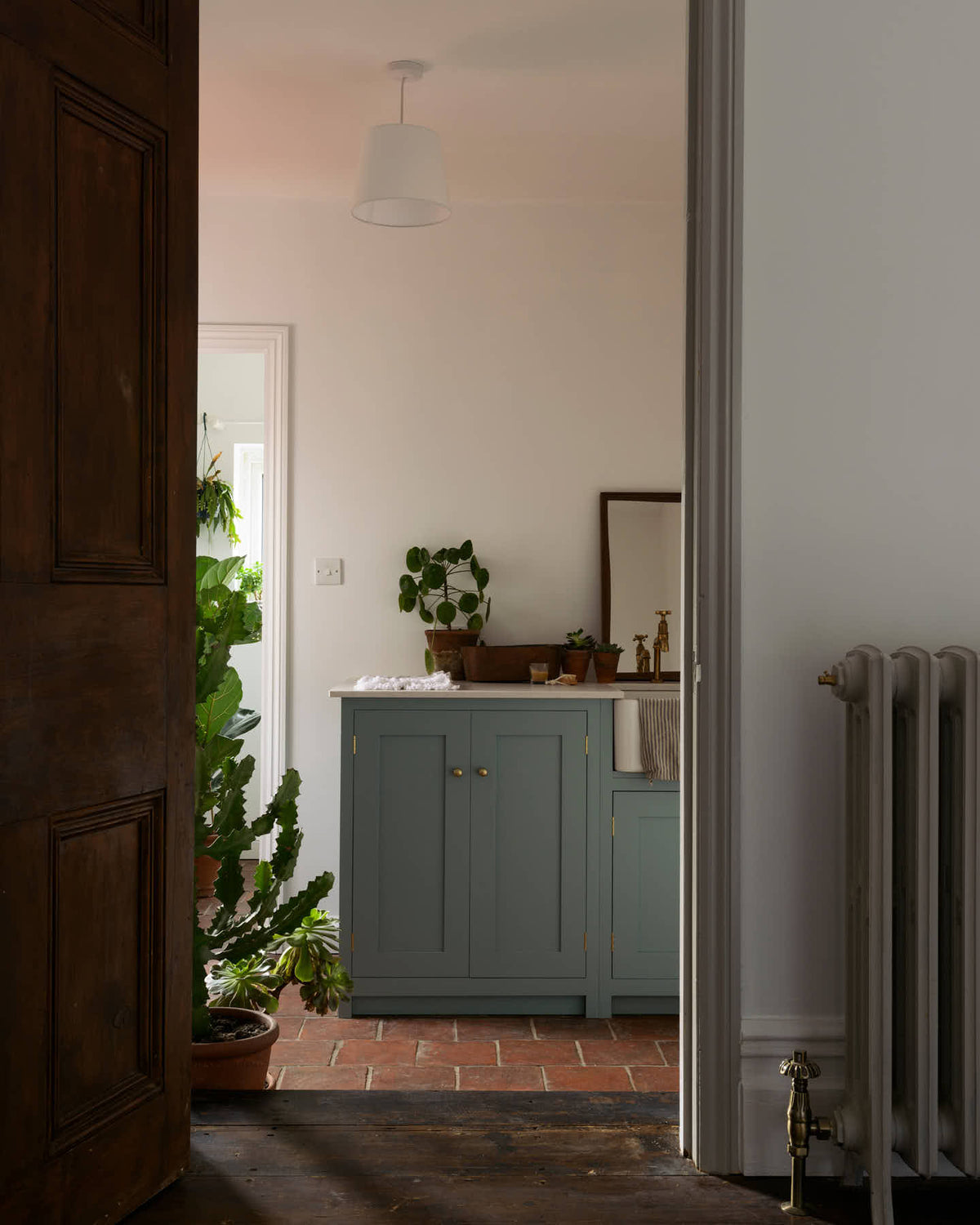 An open door in a well-preserved house with original floorboards reveals a small Trinity Blue scullery room, featuring a Carrara marble countertop, a single Belfast sink with aged brass Mayan taps, and warm terracotta tiles.