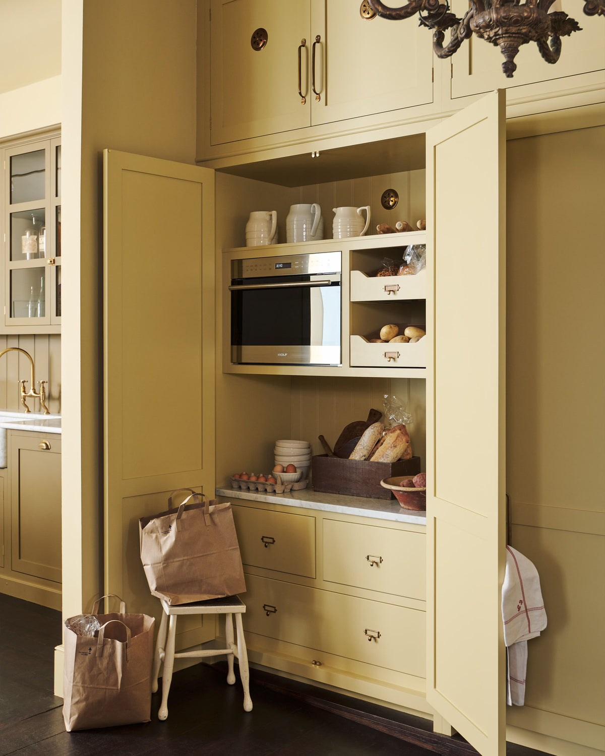 An oblique view of a simple corridor that houses three sets of double cupboards; the left-hand side is open, revealing a pantry cupboard with a microwave. Naturally decorated with a tea towel, artisan ceramics, bread, eggs, potatoes, and an antique chandelier, creating a clean and organised storage space.