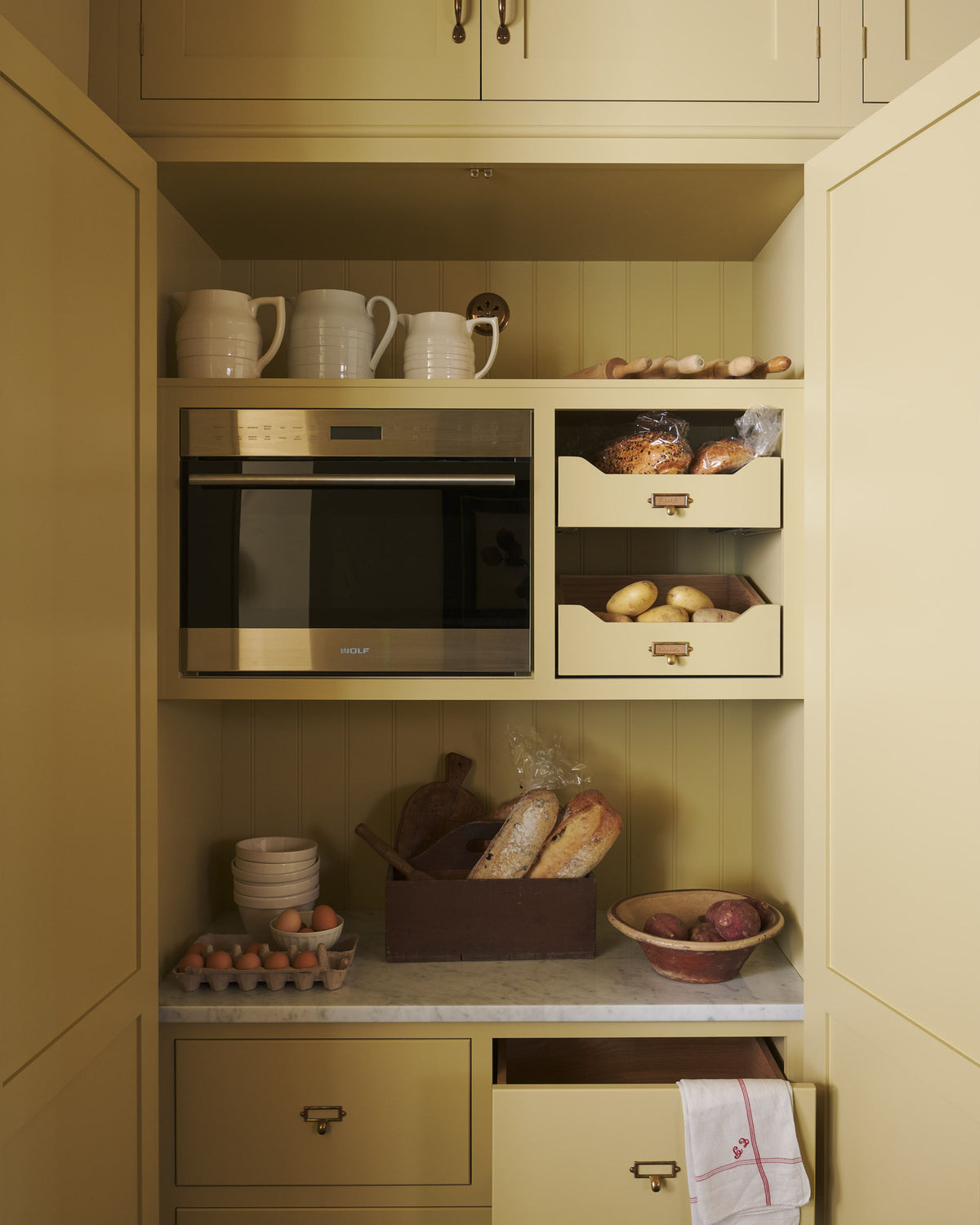 A simple corridor that houses three sets of double cupboards; the left-hand side is open, revealing a pantry cupboard with a fitted microwave, a Carrara marble shelf, and a top cupboard with brass ventilation grilles. Naturally decorated with a little label holders, tea towel, artisan ceramics, bread, eggs, potatoes, and an antique chandelier, creating a clean and organised storage space.