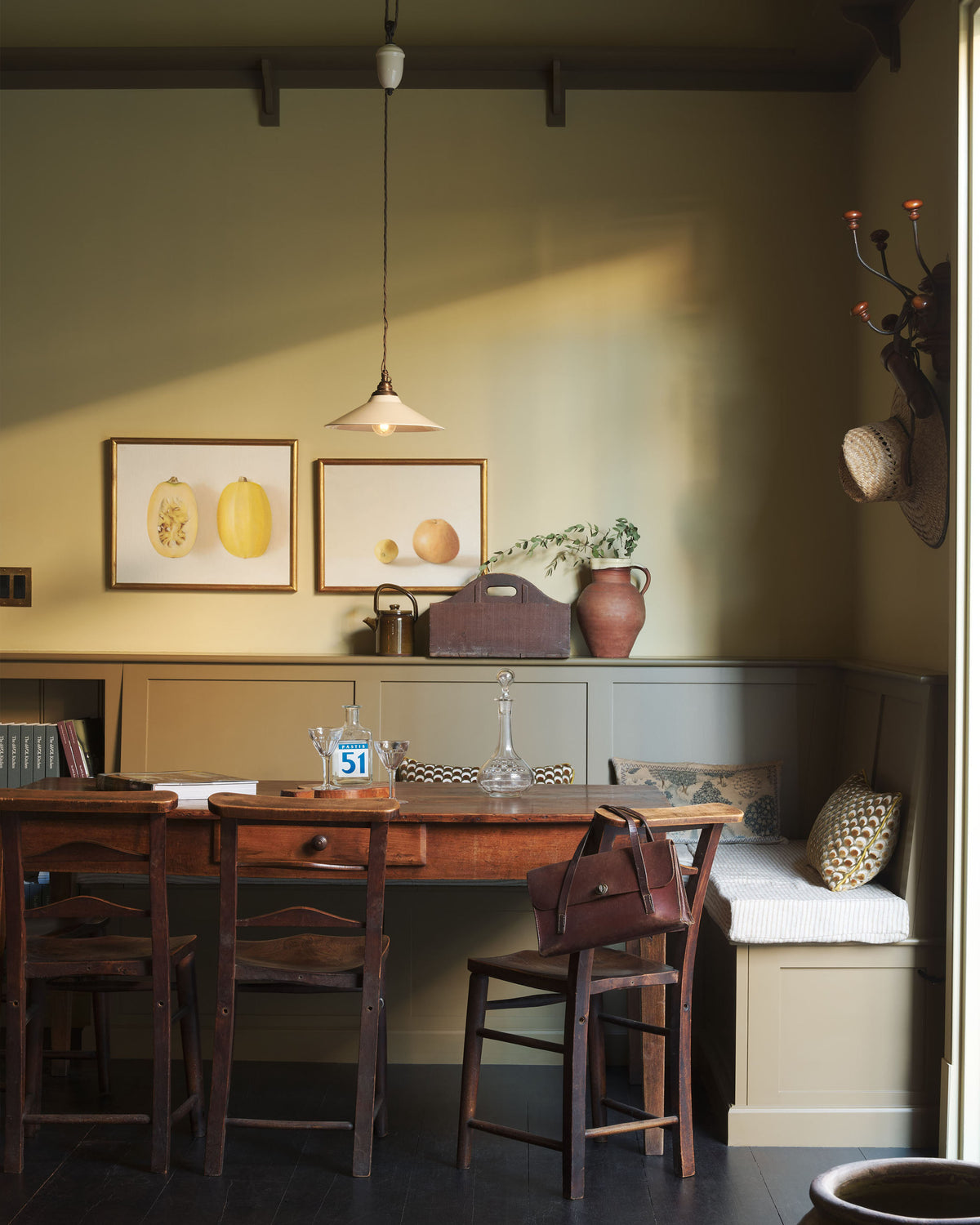 A simple buttery yellow Shaker living space features a Folgate green bespoke bench seat with an open base cabinet, a shelf, a rise and fall pendant light, and a cohesive set of vintage table and chairs. Lovingly adorned with artisan ceramics, paintings, books, a coat rack and cushions, creating a cosy and Mediterranean cafe-inspired social space.