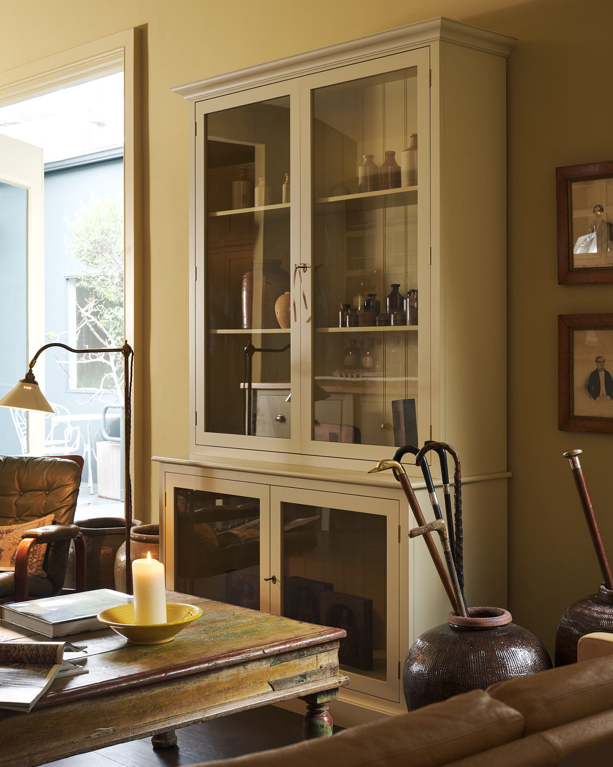 An oblique view of a simple buttery yellow Shaker living space features a Quaker cream curiosity cupboard, a grand tour reading lamp, and a cohesive set of vintage furniture. Lovingly adorned with artisan ceramics, books, and antique canes, creating a humble and mid-century inspired social space.