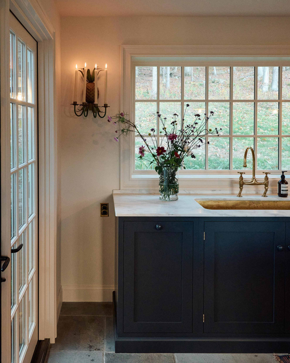 A quiet corner of a cool, spacious Pantry blue Shaker kitchen features a long sink and cooker run with a Carrara marble worktop, and an aged brass ionian tap. Lovingly decorated with a large vase of wild flowers and an antique wall candle holder, it creates a unique and practical kitchen space.
