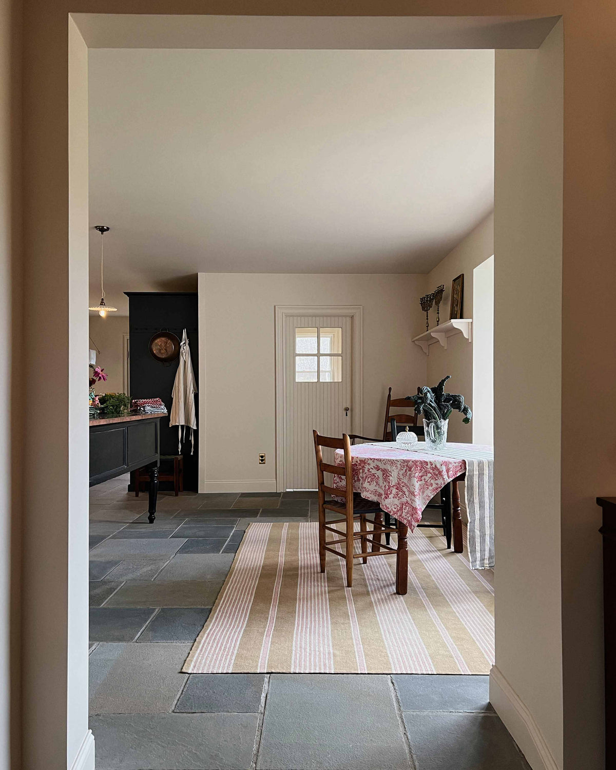 Looking through an open corridor lies a cool, spacious Pantry blue Shaker kitchen featuring a bank of appliance cabinets and a pantry larder. Beside it is a small dining area with antique wooden tables and chairs. Lovingly decorated with a large rug, red detailed cloth, houseplants, and fresh vegetables, it creates a humble, practical kitchen and dining space.