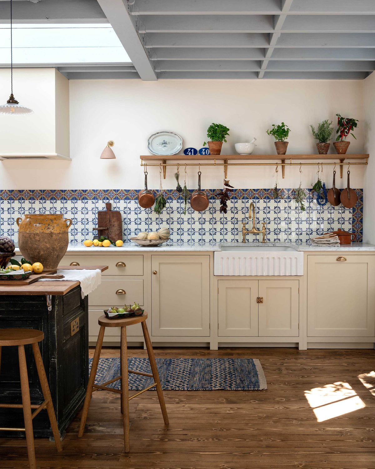 A spacious, Mediterranean-inspired Quaker Cream Shaker kitchen features a long sink and cooker run with a Carrara marble worktop, and a classic oak shelf. Lovingly decorated with a wall of vibrant blue patterned splashback tiles, aged brass hanging rails, Helen’s Stools, green house plants, and fresh produce, it creates a wonderfully social and practical kitchen space.