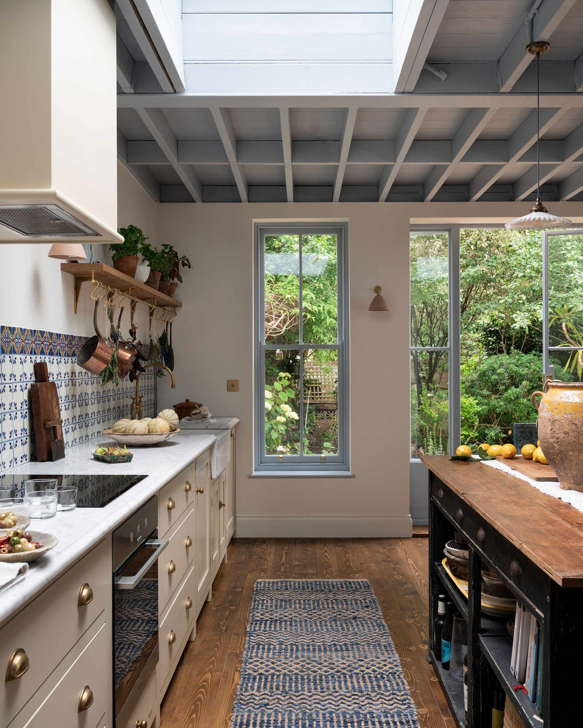 Looking along the galley of the Mediterranean-inspired Quaker Cream Shaker kitchen featuring a long sink and cooker run with a Carrara marble worktop, and a classic oak shelf, it looks through the luscious green garden. Lovingly decorated with a wall of vibrant blue patterned splashback tiles, aged brass hanging rails, green house plants, and fresh produce, it creates a wonderfully social and practical kitchen space.