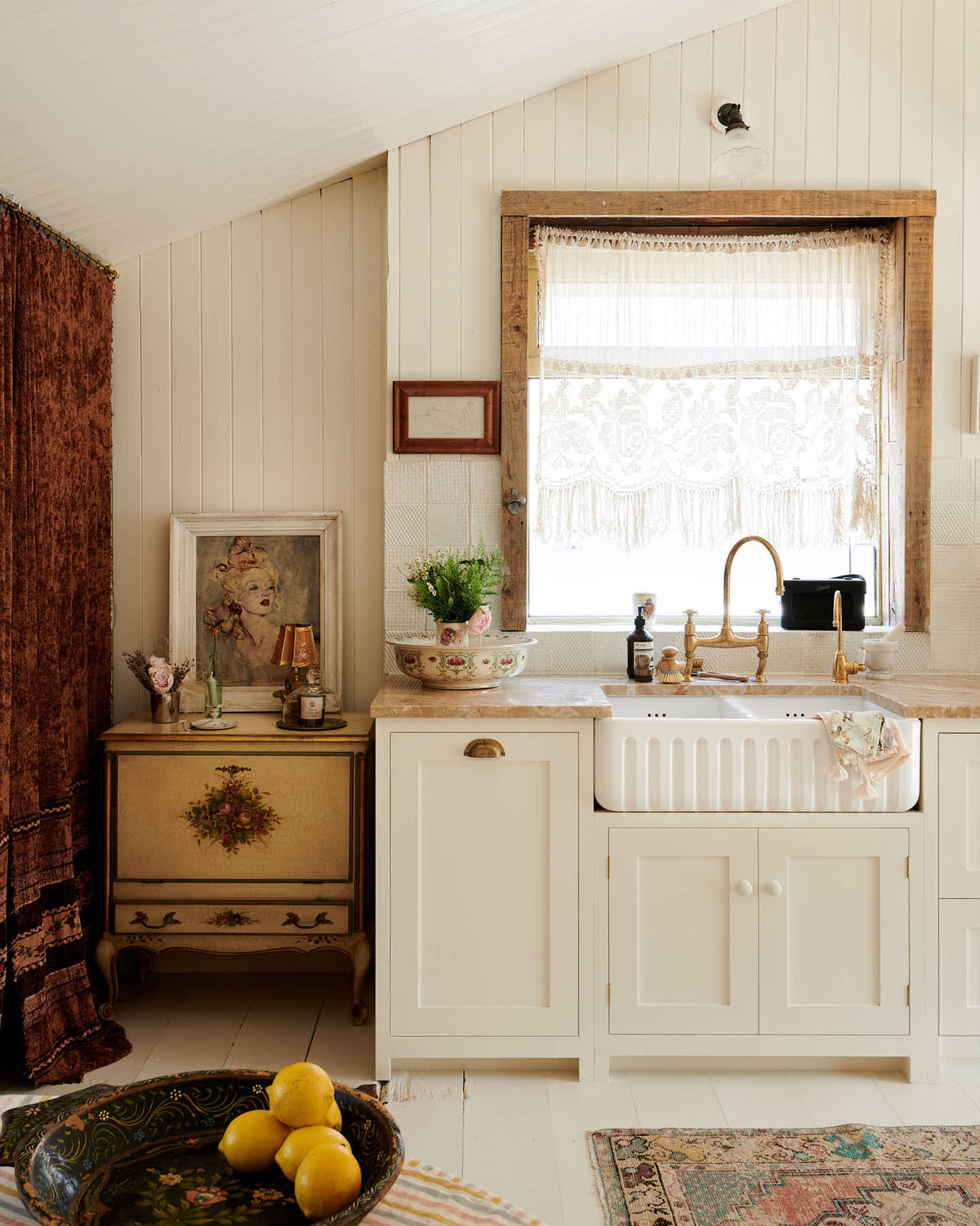 A bohemian kitchen with cream coloured Shaker-style cupboards, floorboards and walls. The room is decorated with an assortment of vintage pieces of furniture, paintings and fabrics.