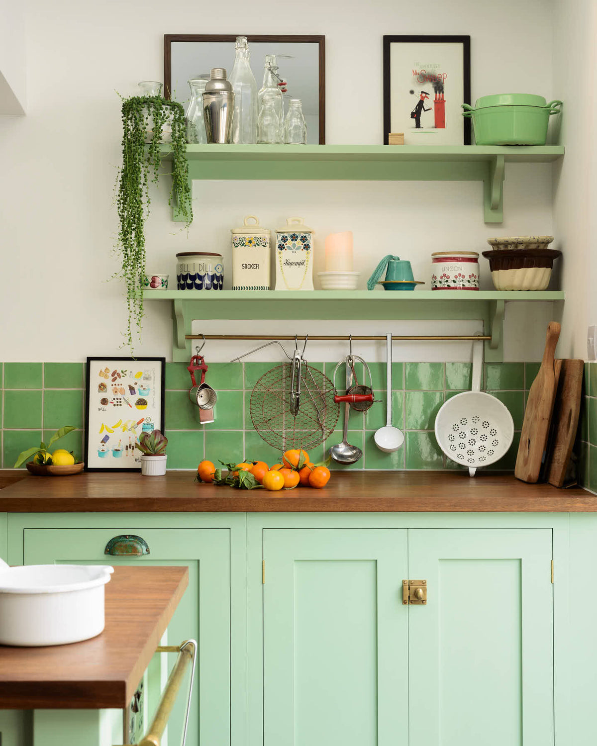 A corner of a pea-green Shaker kitchen features a sink run and a kitchen island with a gas cooker, both topped with a reclaimed iroko school countertop, matching green square splashback tiles, and shelves with brass hanging rails. Beautifully decorated with artisan ceramics, lemons, oranges, and a string-of-pearls plant, it creates a friendly and welcoming kitchen.