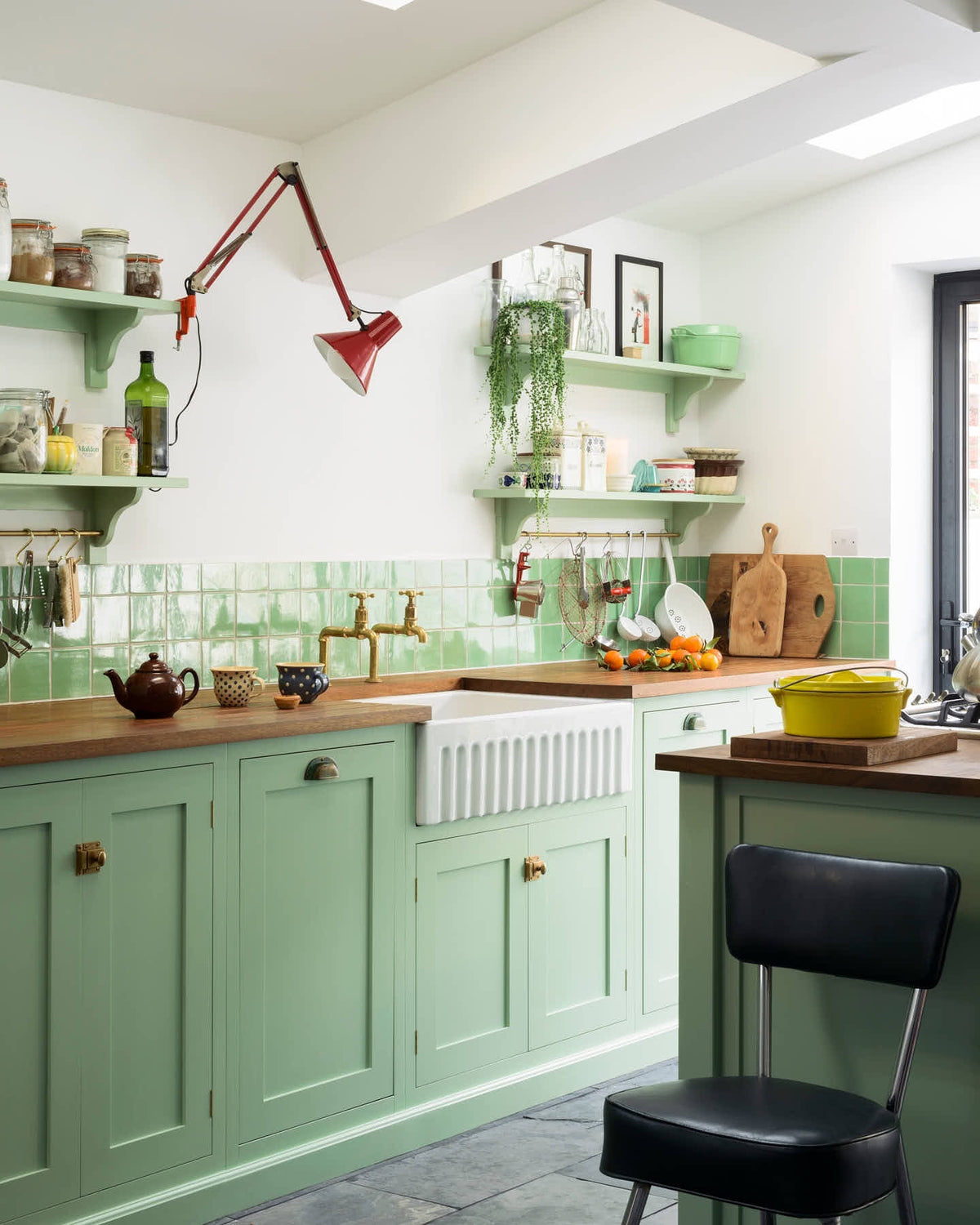 An oblique view of a vibrant pea-green Shaker kitchen features a sink run and a kitchen island with a gas cooker, both topped with a reclaimed iroko school countertop, matching green square splashback tiles, shelves with brass hanging rails, and a double oven cabinet with drawers. Beautifully decorated with artisan ceramics, dried ingredients in a glass jar, oranges, and a clamped red lamp, creates a friendly and welcoming kitchen.