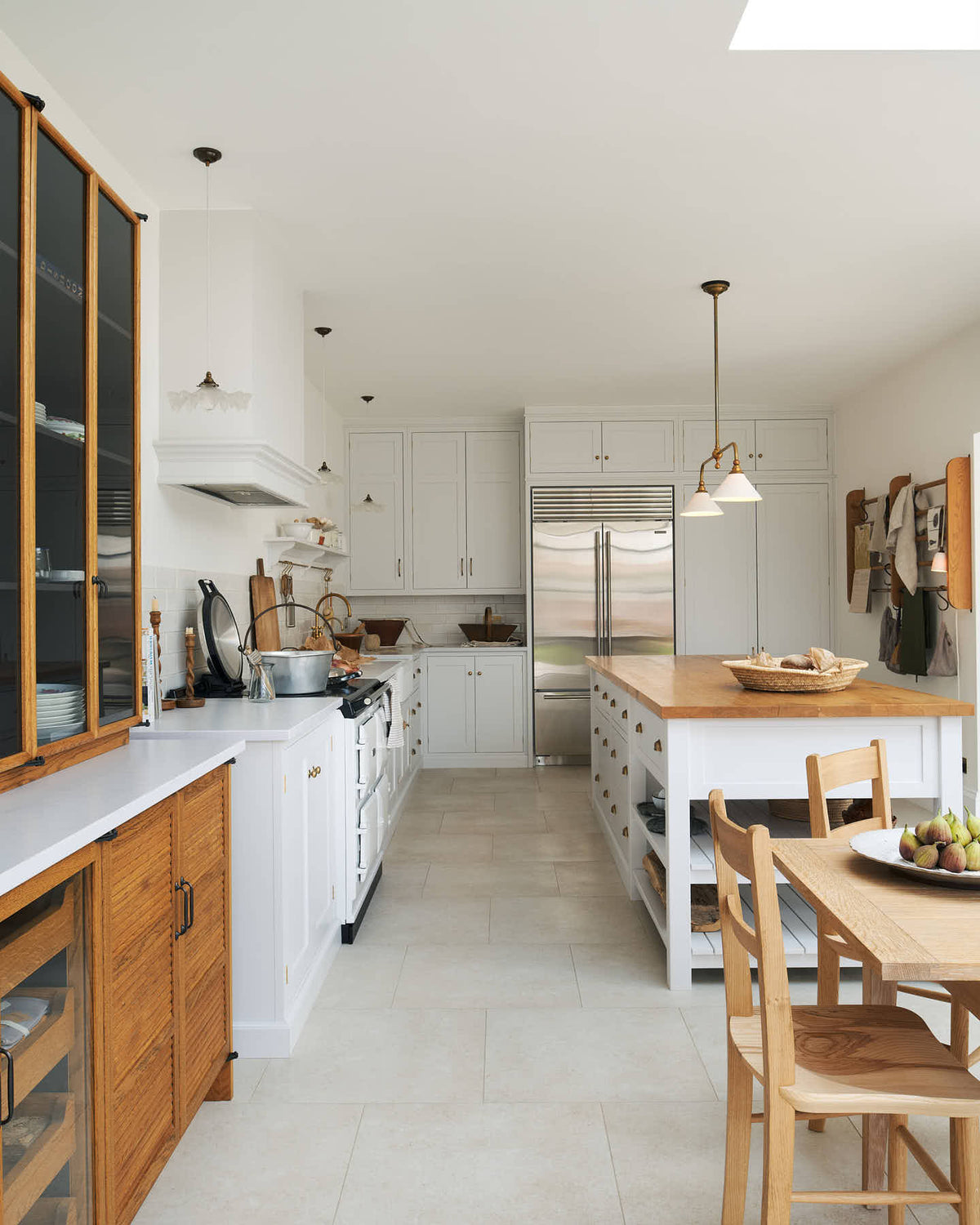 Looking down at a minimalist Damask off-white shaker kitchen with a solid oak island and Carrara marble worktops for the sink and cooker. The sink and cooker runs feature crackle metro splashback tiles. A shaker table with oak chairs and glazed countertop cupboards complete the space, accented by warm browns and golds, from wooden chopping boards to the aged brass Heirloom gaselier light.