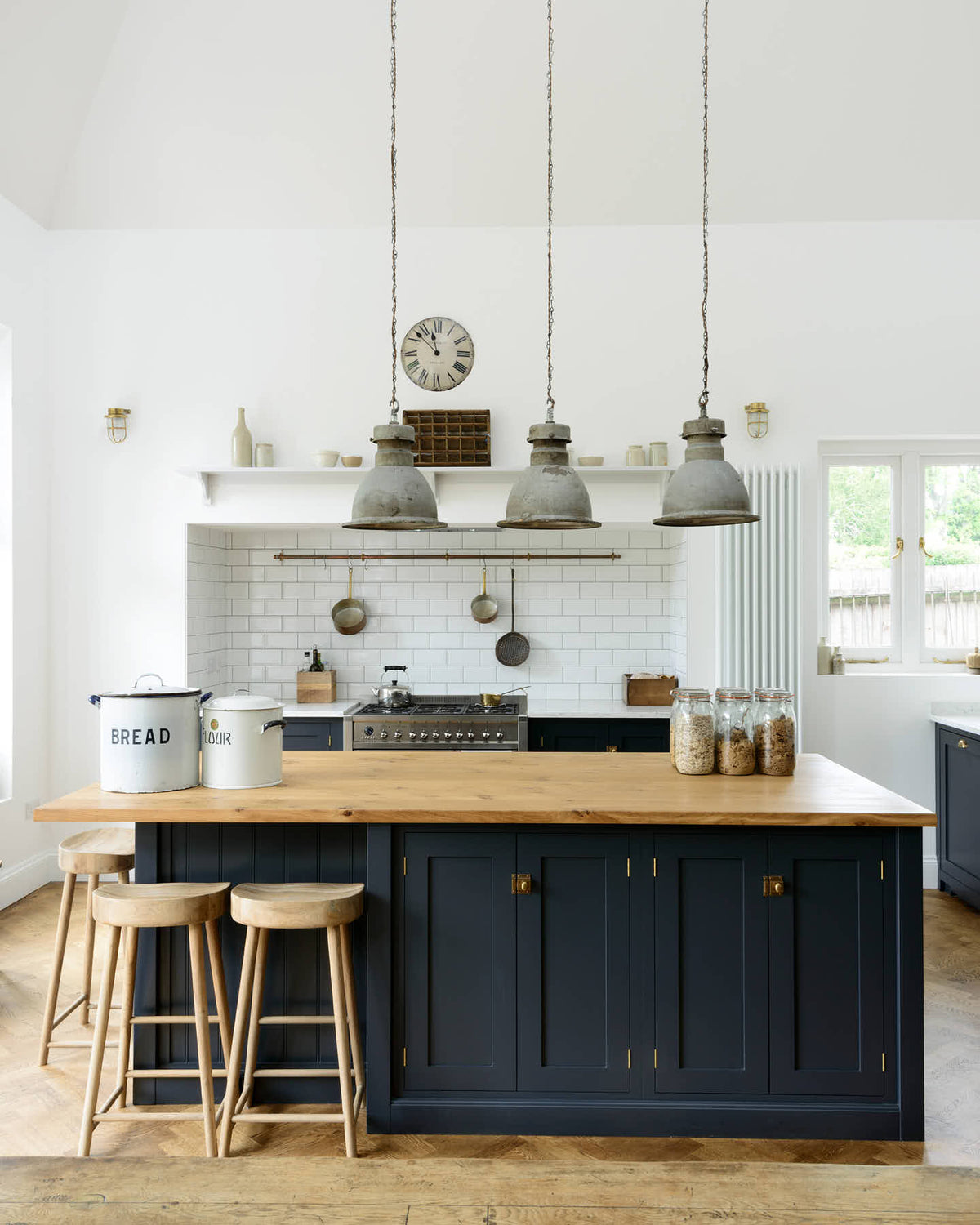 A tall pantry-blue shaker kitchen features a sink and cooker run with a Silestone worktop, complemented by a large kitchen island topped with solid oak and glazed cupboards. The cooker run sits in an alcove with white splashback tiles, topped by a shaker shelf displaying assorted ceramic and wooden bric-a-bracs. Rusty metal hanging lights in the centre add an industrial touch to the space.