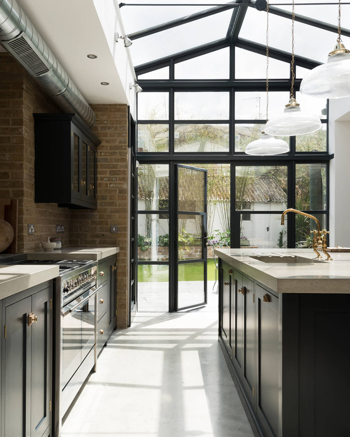 A bright and spacious pantry-blue Shaker kitchen under a wall-to-ceiling skylight features a long cooker run with wall cabinets, full-height cupboards, a kitchen island with a sink, a concrete worktop, and a tall bookshelf. Adorned with glass pendant lights, a muted pestel and mortar set, and chopping boards, it creates a warm and social space.