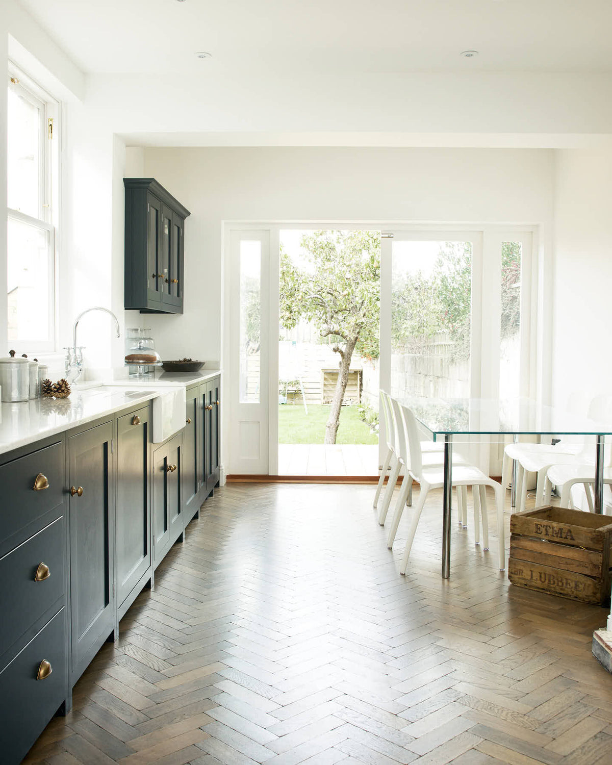 An oblique view of a bright and simple pantry-blue Shaker galley kitchen features a long sink and cooker run with a double Belfast sink, an antique silver Ionian tap, and glazed wall cupboards. Beautifully decorated with fundamental kitchen equipment such as a silver toaster, matching silver coffee, tea, and sugar containers, a glass cake stand and dome, and reclaimed wooden parquet, it creates a stylish and effortless space.