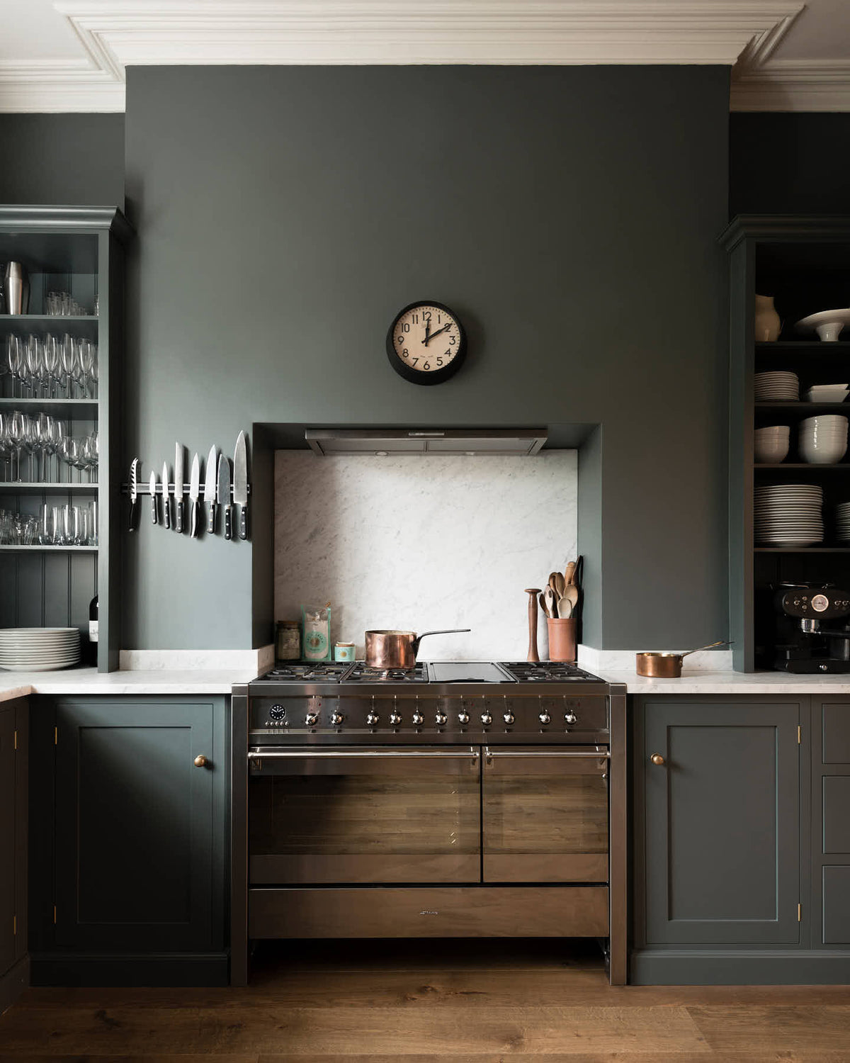 A charcoal-black, classic shaker kitchen features a U-shaped sink and cooker run with Carrara marble worktop and splashback, a double Belfast sink, and a silver gas stove. Beautifully decorated with a magnetic kitchen knife holder, a steampunk wall light, a steel weight, and branches of purple and green flowers, the family kitchen strikes a wonderful balance between modern style and timeless character.