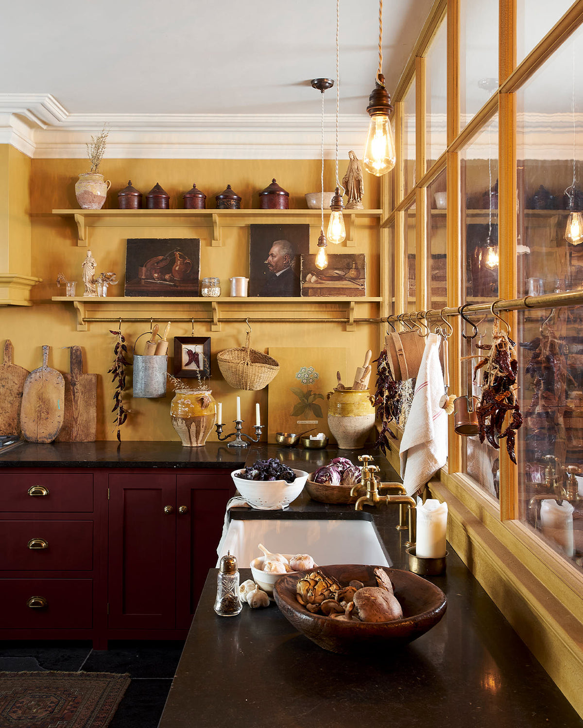 A busy and vibrant Refectory Red Shaker kitchen features an L-shaped sink and cooker run with a Belgian blue limestone worktop, shelves with brass rails, and a classic Shaker canopy. Adorned with vintage items, beloved ceramics, a still-life painting, wooden chopping boards, and dried chillies, creates a bright and wonderfully social kitchen space.