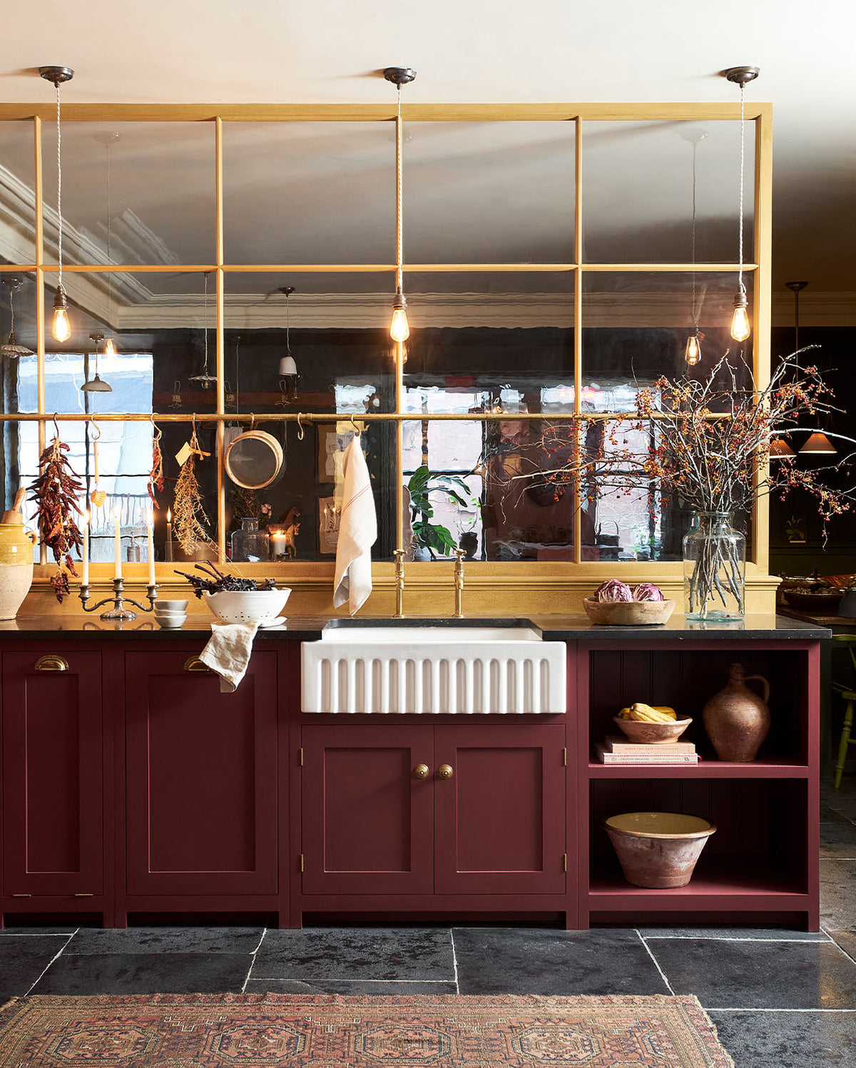 A busy and vibrant Refectory Red Shaker kitchen features an L-shaped sink and cooker run with a Belgian blue limestone worktop, a single fluted Belfast sink, and aged brass Mayan taps. Adorned with vintage items, beloved ceramics, still-life paintings, dried chillies, red lettuce, and an arrangement of berry branches, creates a bright and wonderfully social kitchen space.