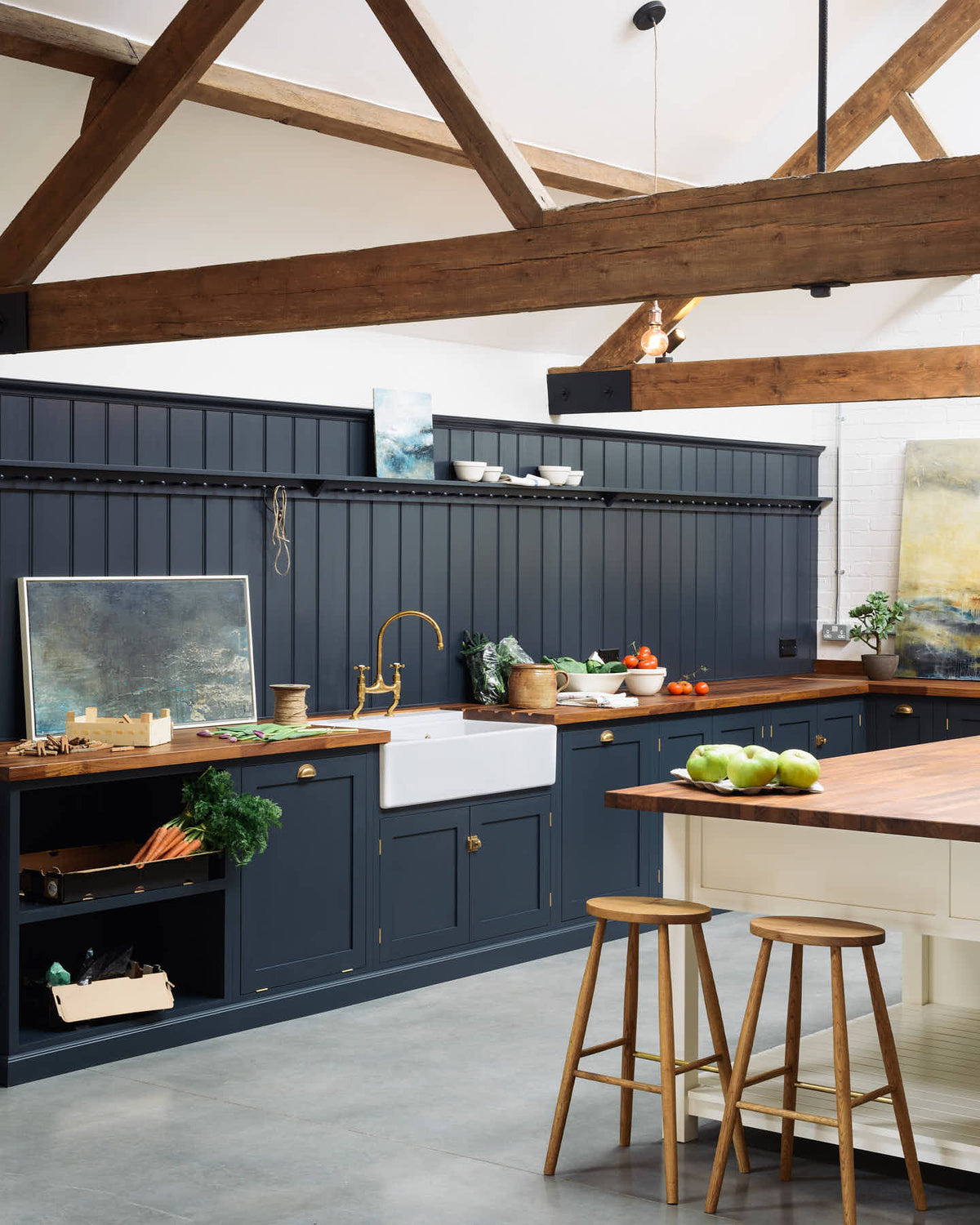 An oblique view of a large, renovated cattle shed has been transformed into a spacious pantry-blue Shaker kitchen, featuring an L-shaped cooker and sink run, along with a large white linen kitchen island topped with a stave hardwood worktop. Beautifully decorated with an aged brass Ionian tap, Helen stools, lucious green vegetables, vintaged artisan ceramics, lilac tulips and abstract paintings.