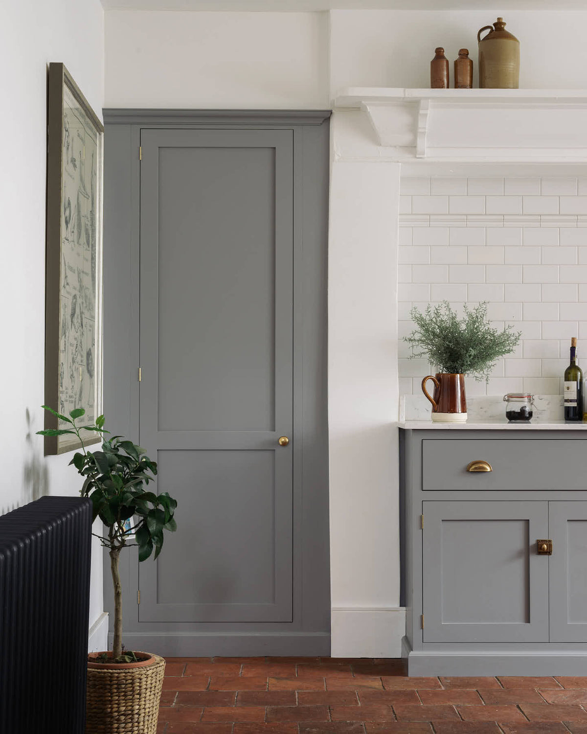 A closer look at the shaker kitchen’s full-height pantry door and the edge of a simple, modern cooker run, finished with white porcelain splashback tiles. Cupboards in mid-grey, terracotta tile flooring, and crisp white walls. Styled with a woven basket plant, a framed vintage illustration, and handcrafted ceramic bowls, jugs, and mugs.