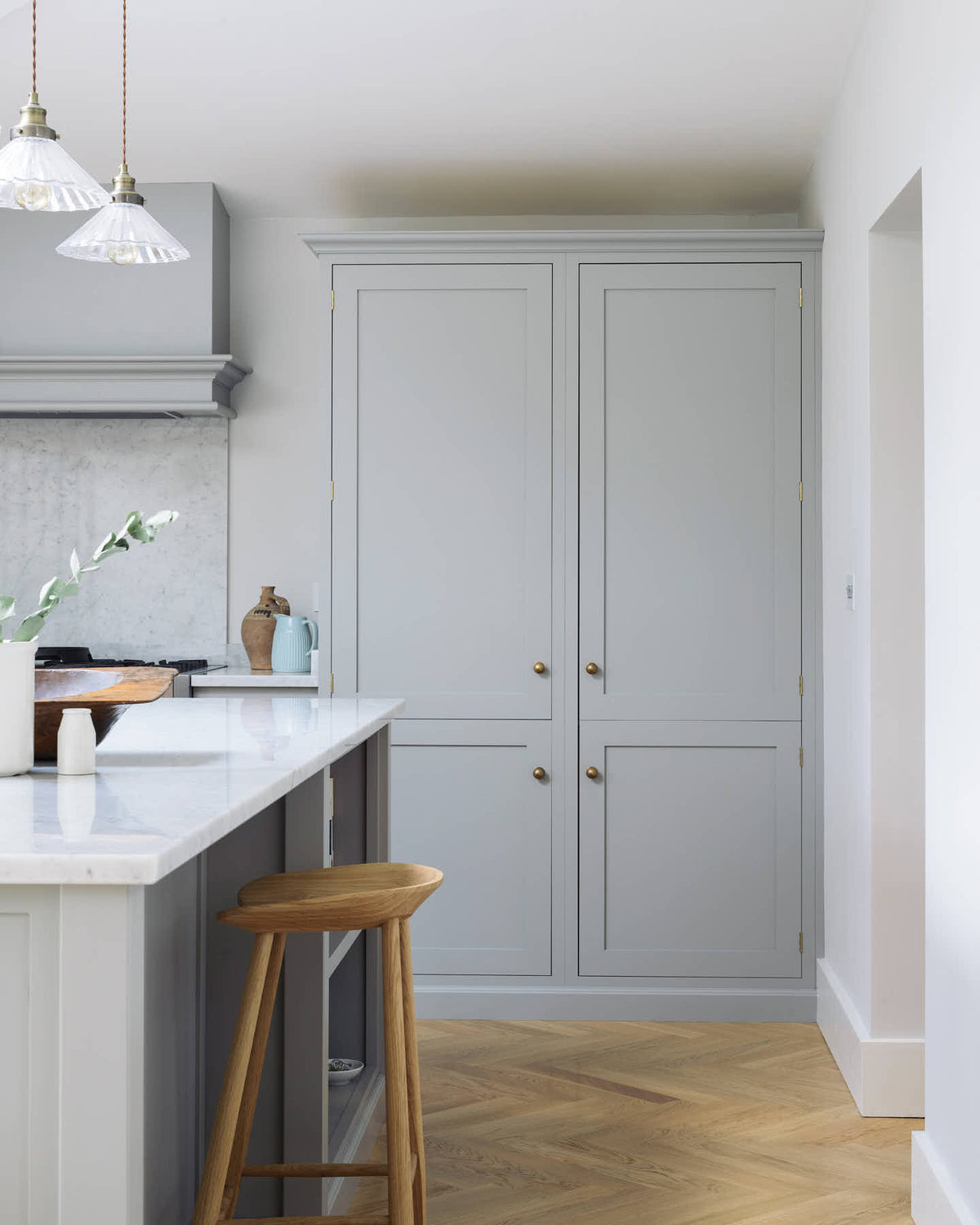 A corner of a spacious and contemporary stone grey Shaker kitchen features an L-shaped sink and cooker run with a Carrara worktop and splashback, a canopy, a bank of full-height cabinets, and a kitchen island. Elegantly decorated with glass-frilled pendant lights, artisan ceramics, and a deVOL bum stool, it creates a cool and inviting space.