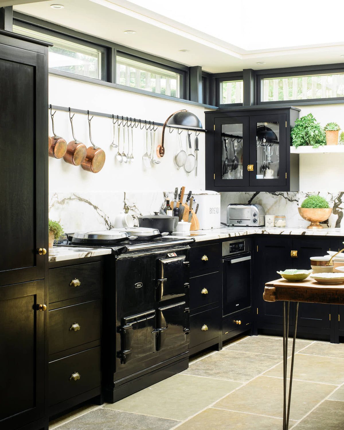 An angled view of a Painter’s Black shake kitchen with an L-shaped layout featuring a cooker and a sink. The worktop and splashback are crafted from San Simone quartzite. The design includes an overhead glazed cupboard and a tall storage cupboard, along with an aged brass hanging rail. The space is illuminated by a glass skylight, enhancing its overall brightness.