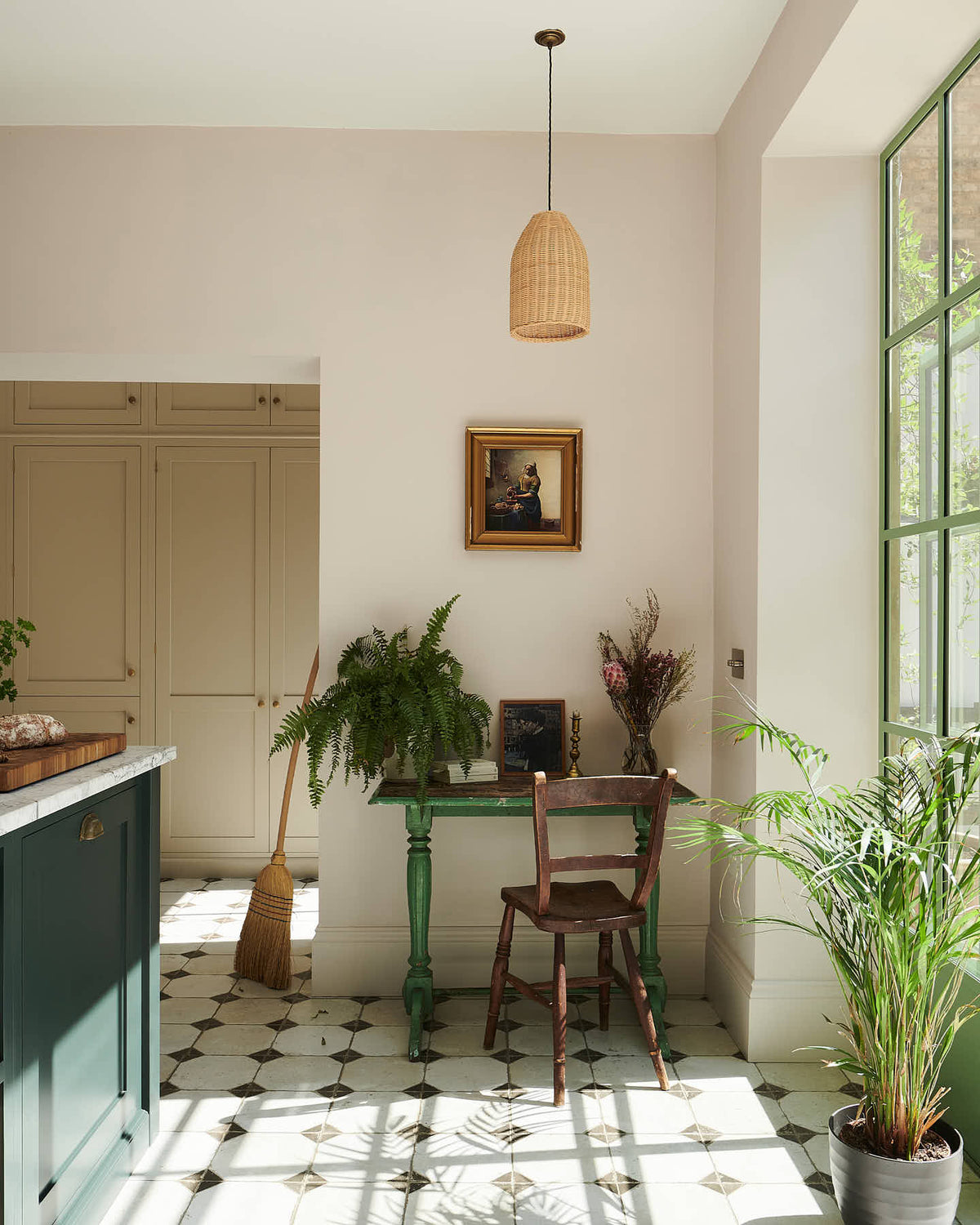 A cosy corner of a Bakehouse Green shaker kitchen features a vintage green wooden desk, a brown chair, and a small painting in a golden frame. This nook also showcases a section of the kitchen island with a Carrara marble worktop, overlooking the pathway to the spacious Mushroom shaker storage and pantry.