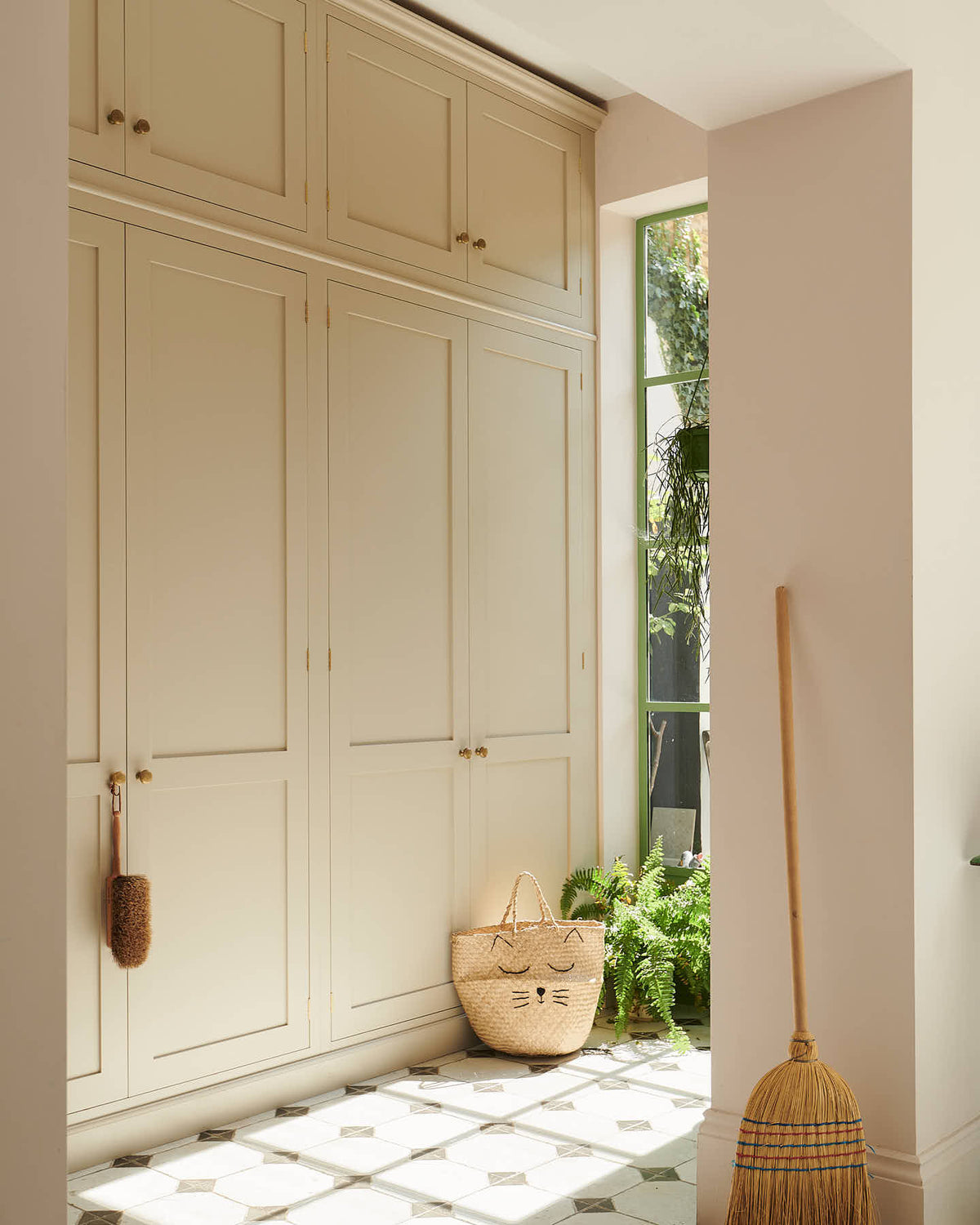 An angled view of the pathway to the spacious, floor-to-ceiling Mushroom shaker storage and pantry. The space features a reclaimed octagonal marble-tiled floor adorned with handmade accessories like a woven basket, broom, and wooden scrubbing brush.