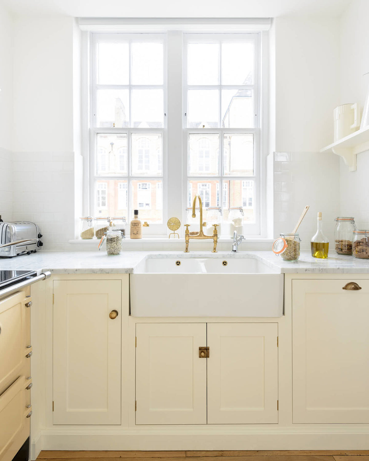 The contemporary living room opens to a small yet functional kitchen with a white shaker design and an L-shaped layout, featuring a cooker and sink with Carrara marble tops. The pale wooden flooring, aged brass handles, and cream gas stove add warmth to this bright kitchen.