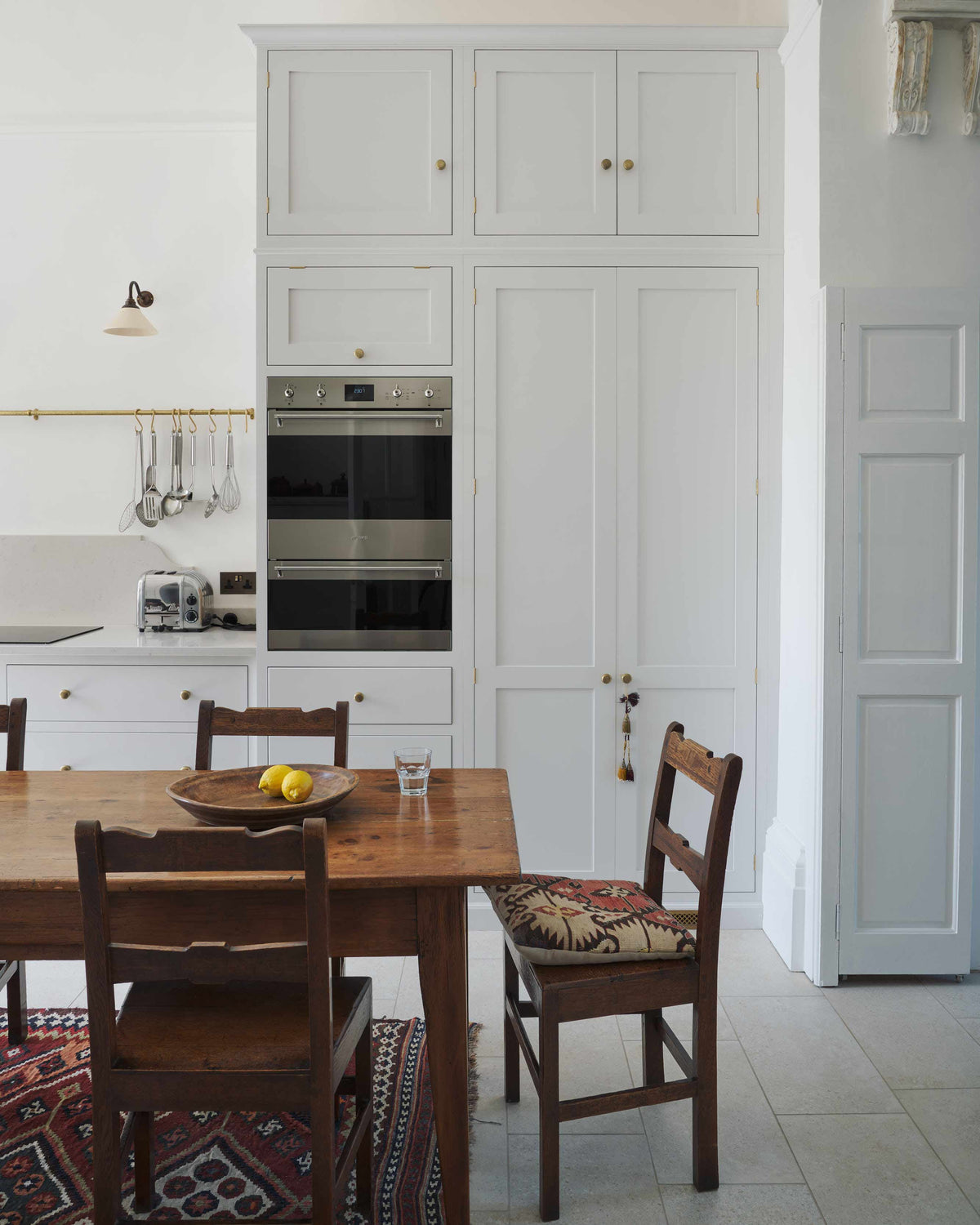 A quiet corner of a tall yet simple Shaker kitchen featuring a small bank of larder. Lovingly decorated with antique furniture, a vibrant red decorative rug, and a variety of handmade charms.